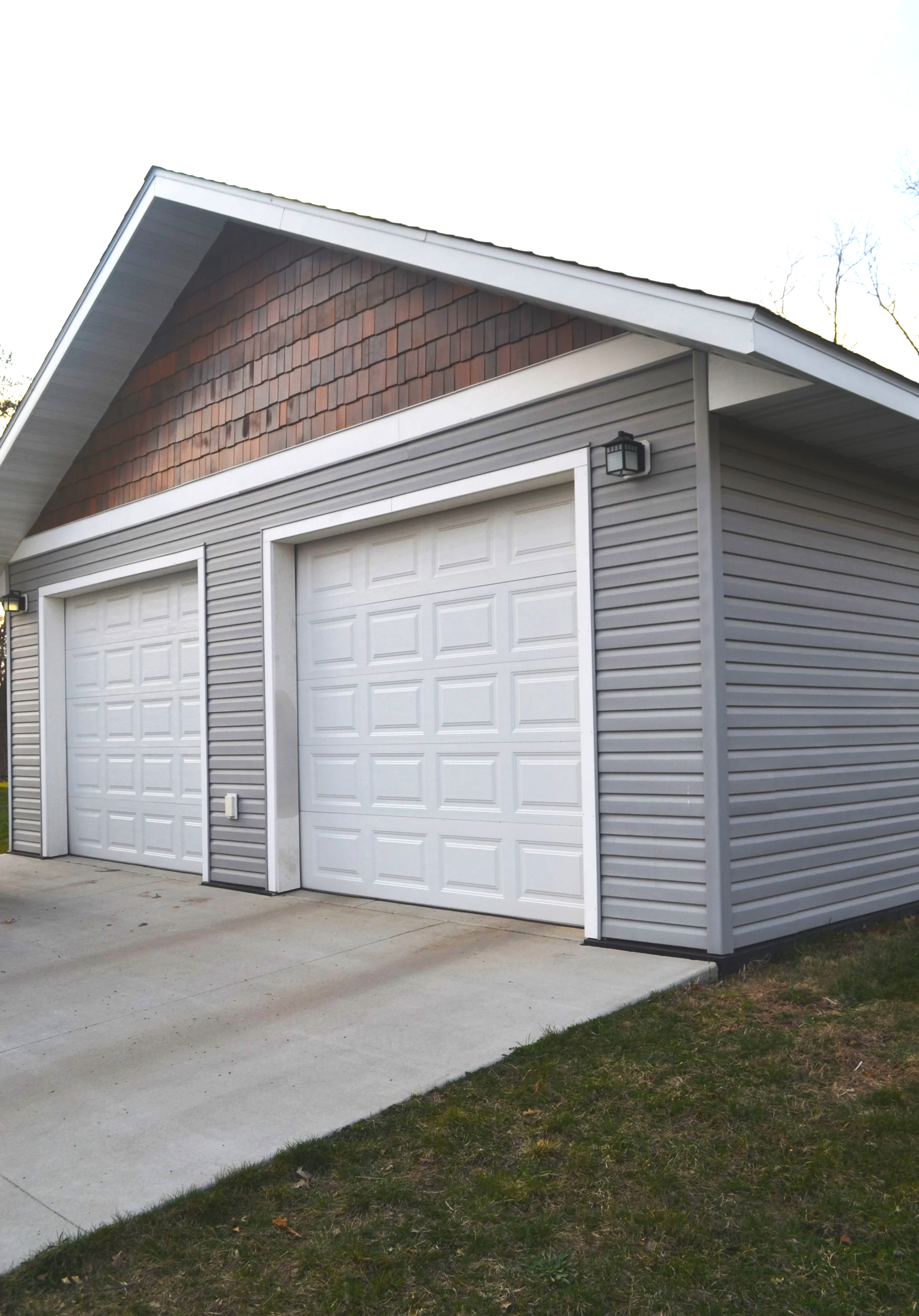 A detached garage with two white doors, gray vinyl siding, and a small outdoor light fixture above the right door, set on a concrete driveway with a grassy yard.