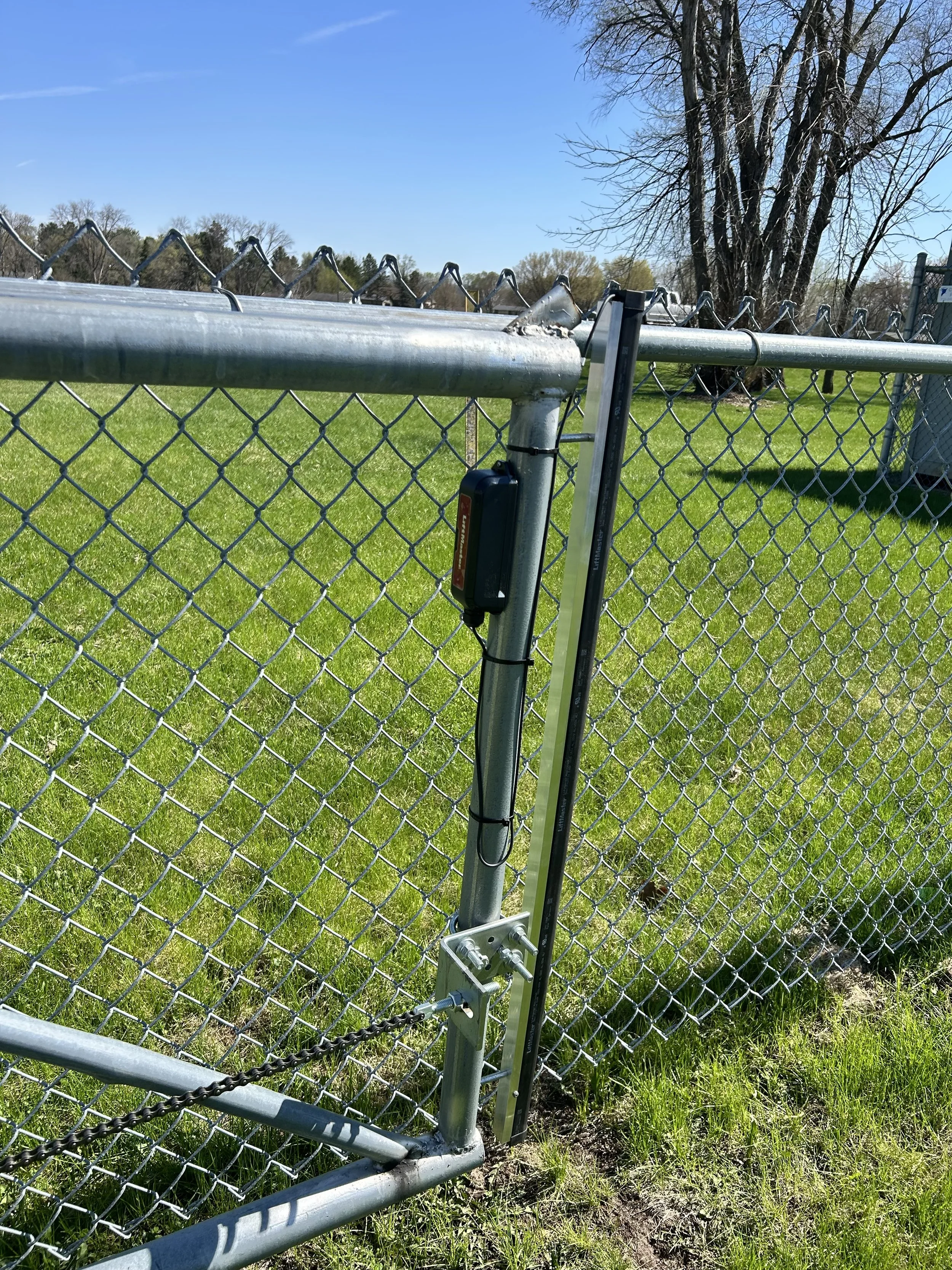 Close-up of a metal chain-link fence gate with a lock and latch, set in a grassy outdoor area with trees and a blue sky in the background.