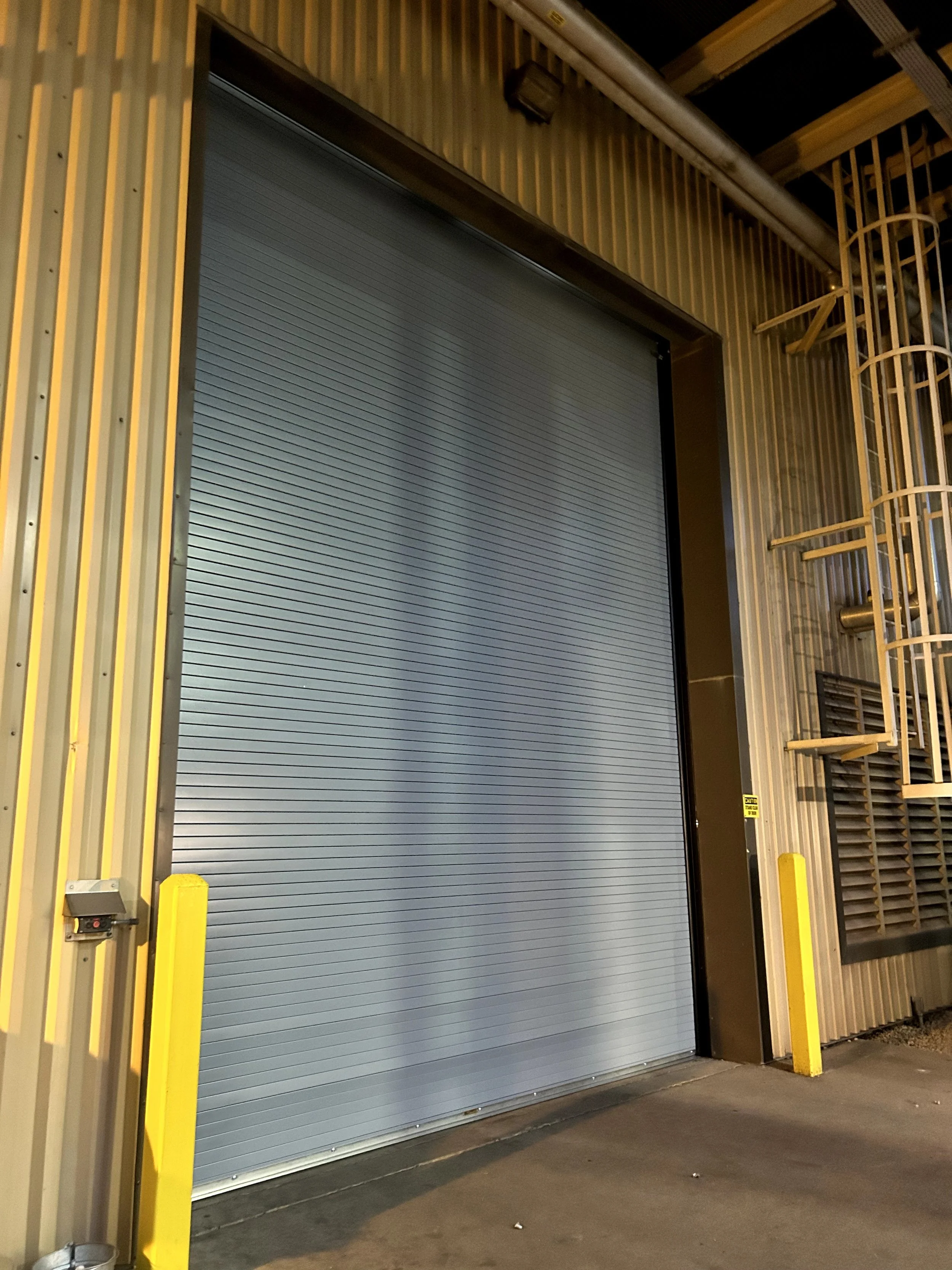 A closed gray industrial roll-up garage door in a warehouse or industrial facility, with yellow safety bollards on either side and a metal siding exterior.