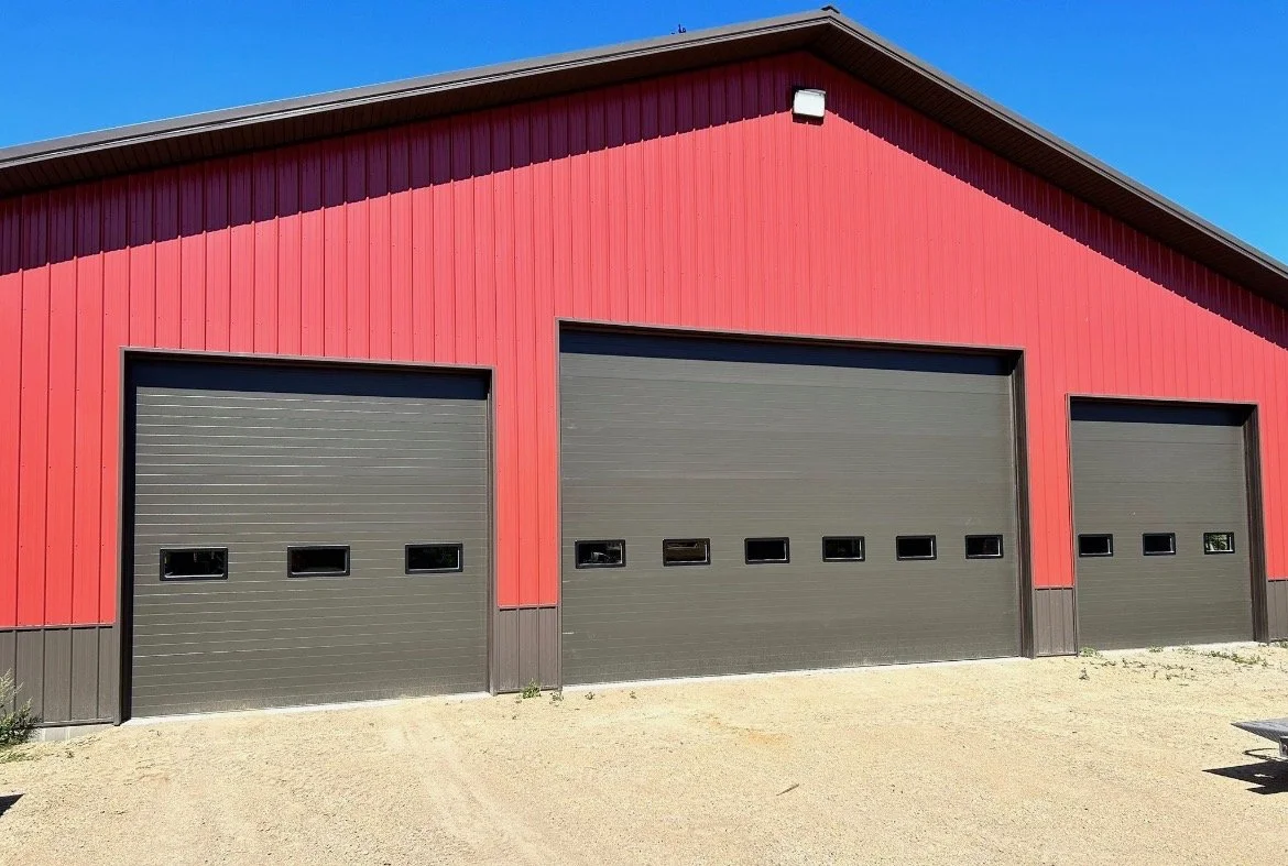 Red barn-style garage with three black roll-up doors, each with small windows, on a sandy driveway under a clear blue sky.