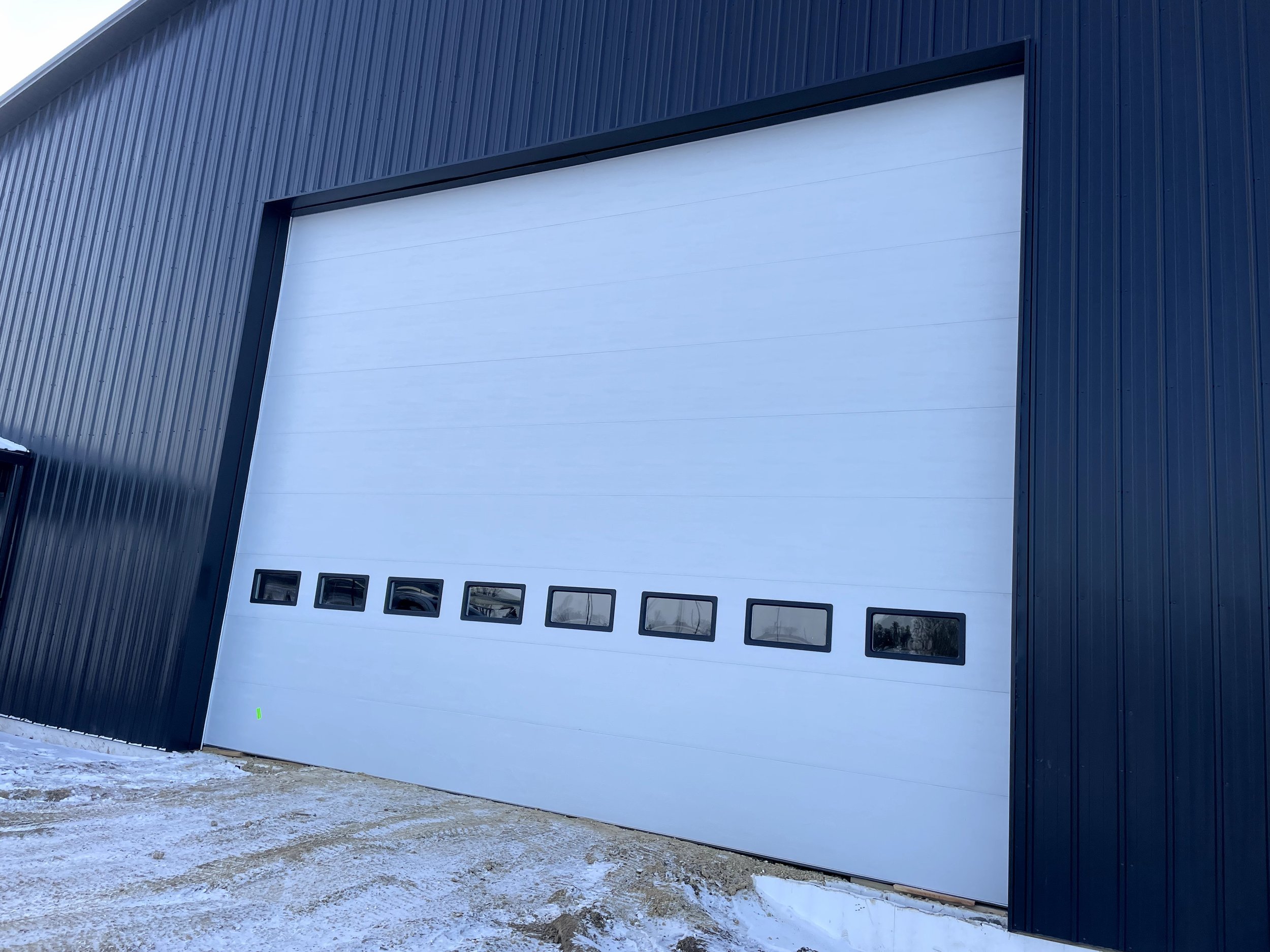 A modern commercial building with a black metal exterior, featuring a large white sectional garage door with small windows near the bottom, and a small patch of snow and dirt in front.