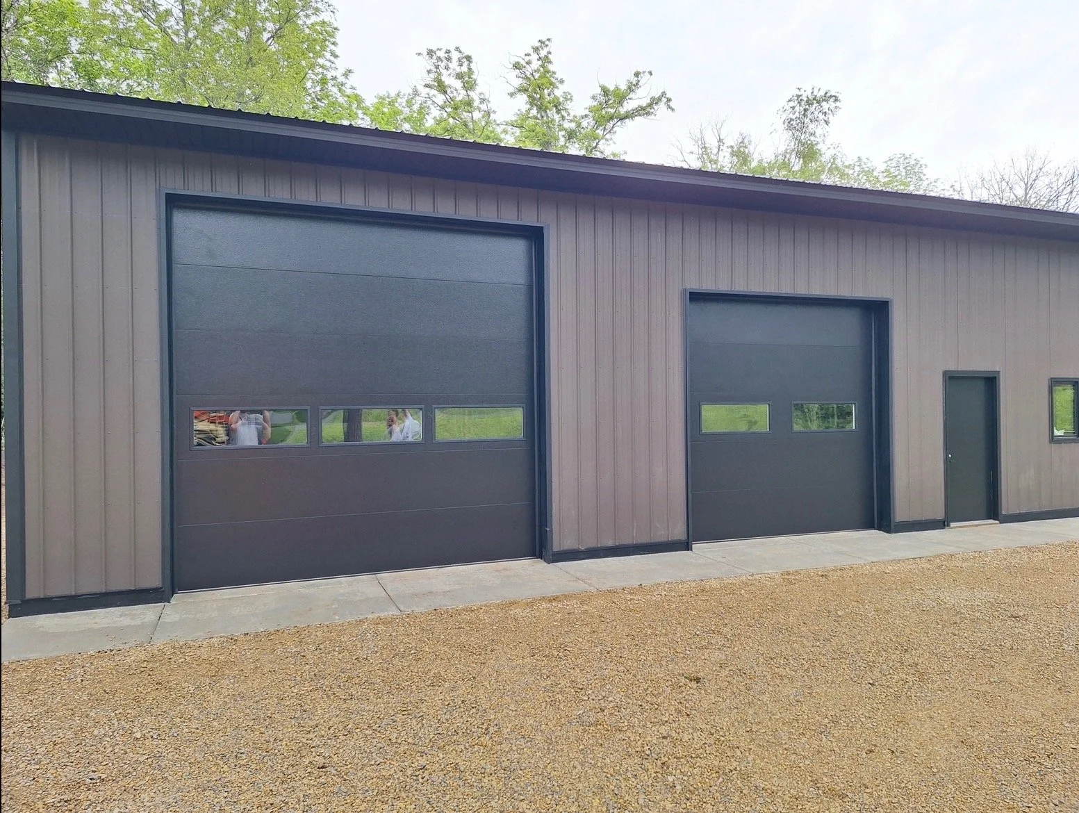 A large modern building with two black garage doors, a black pedestrian door, and small windows, situated on a gravel lot with trees in the background.
