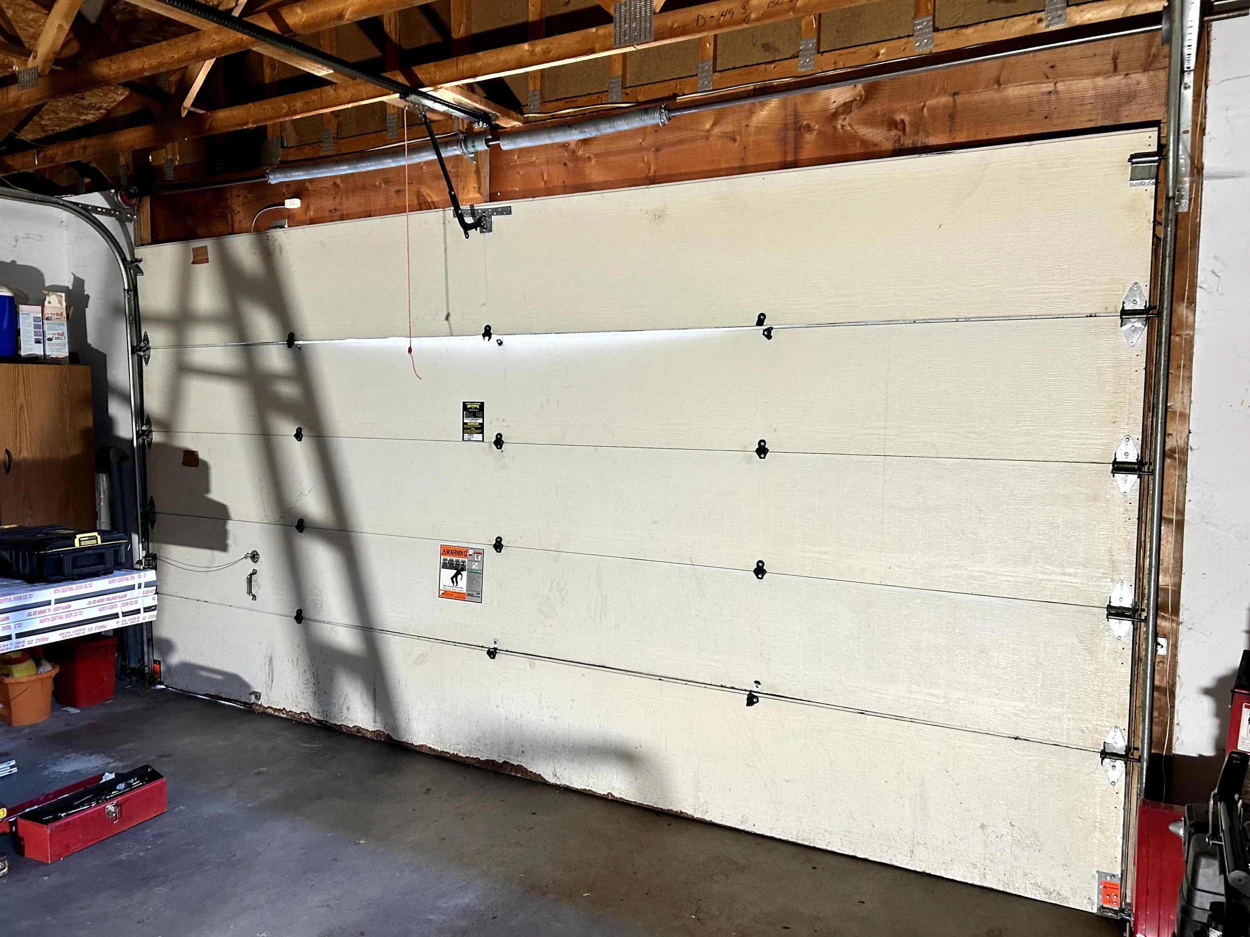 Empty garage with white sectional roll-up door, overhead garage door opener system, and exposed wooden ceiling beams.