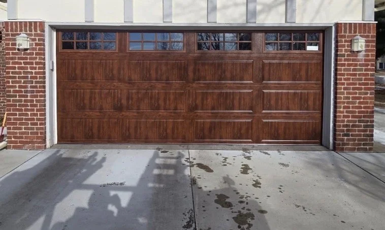 A closed brown wooden garage door with small windows at the top, flanked by brick and white siding, with a concrete driveway in front and water stains visible on the ground.