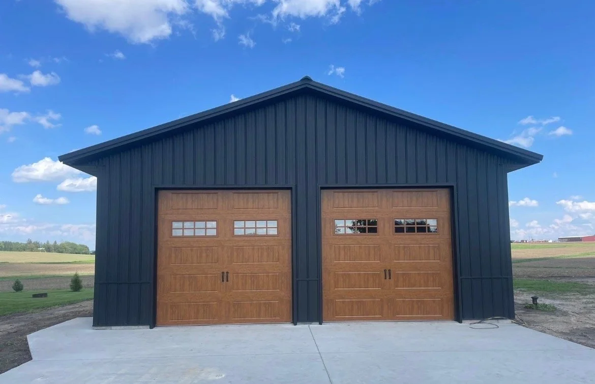 A large black metal garage with two wooden doors, each having a row of small windows at the top, set on a concrete driveway in a rural area under a blue sky with scattered clouds.