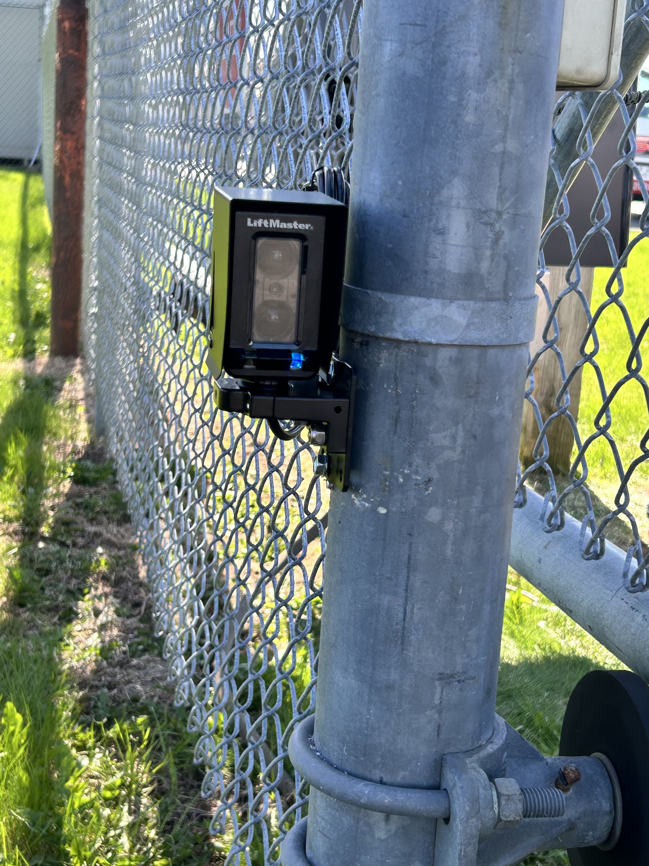 Close-up of a gray metal fire hydrant attached to a chain-link fence during daytime with grass and a street in the background.
