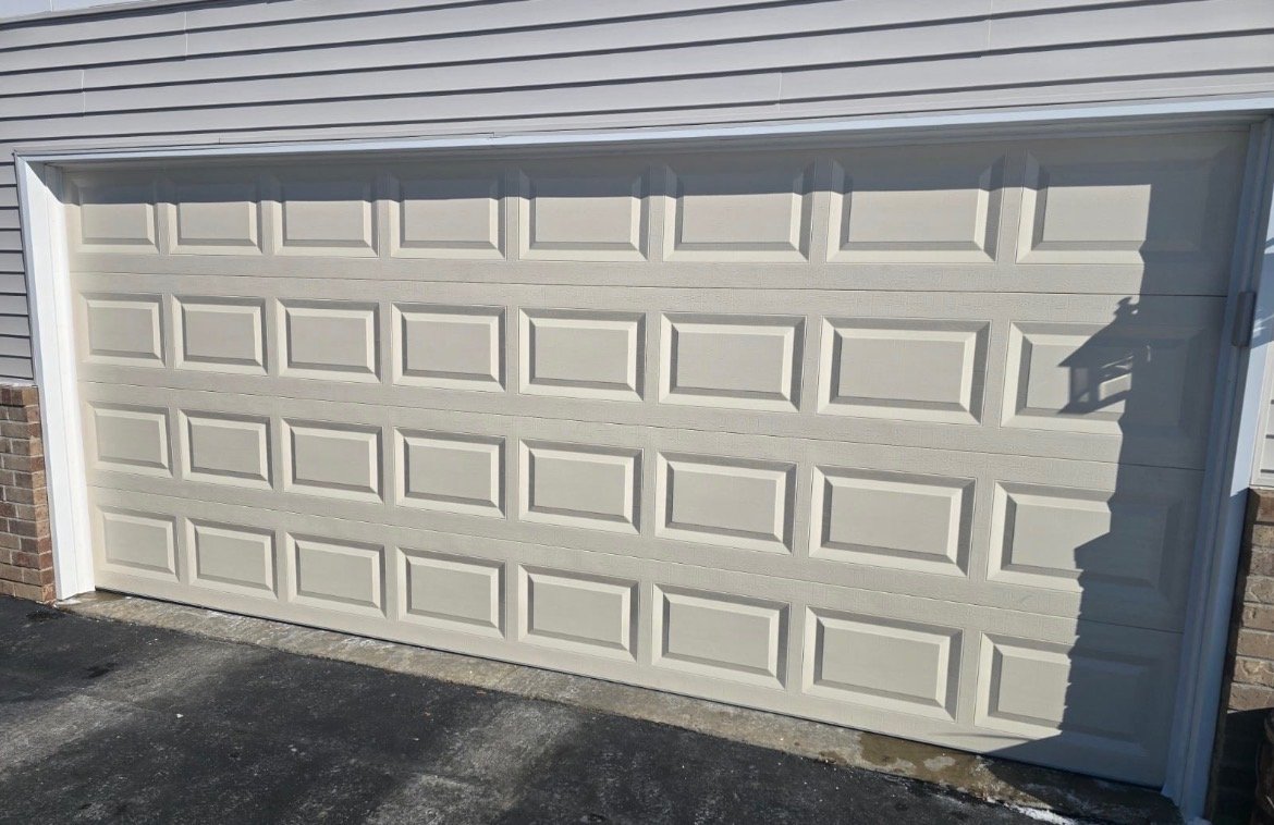 White sectional garage door with a patterned panel design, installed as part of a house exterior with brick and siding, with shadows cast on the door.