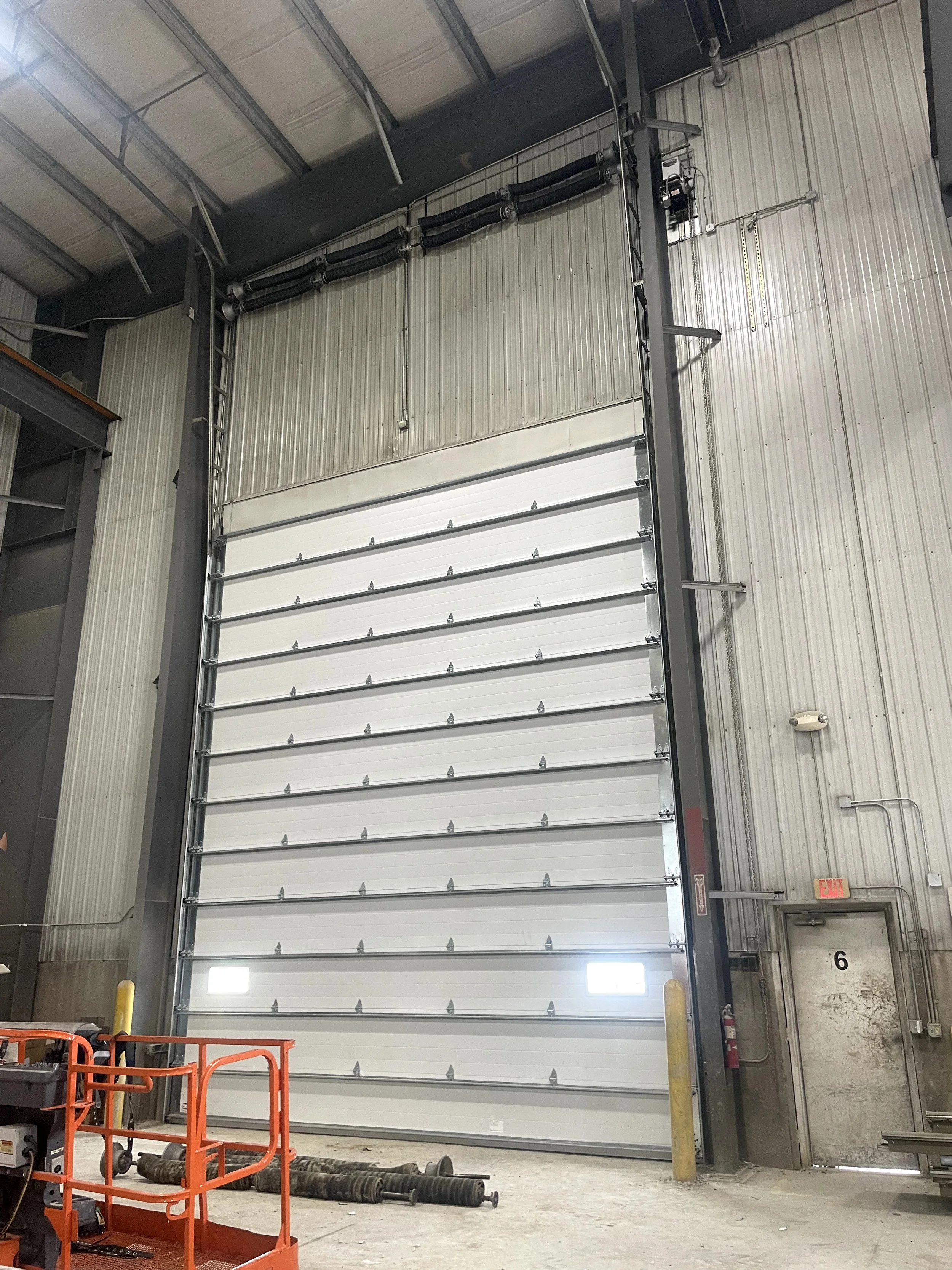 Industrial warehouse interior with a large white rolling door, concrete floor, and equipment such as orange carts and pipes, with yellow safety posts near the door, and an exit sign above a small room.
