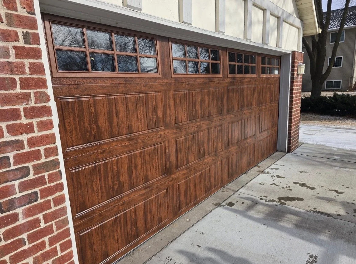 A wooden garage door with multiple rectangular panels and small window panes at the top, set against a brick wall and a concrete driveway.