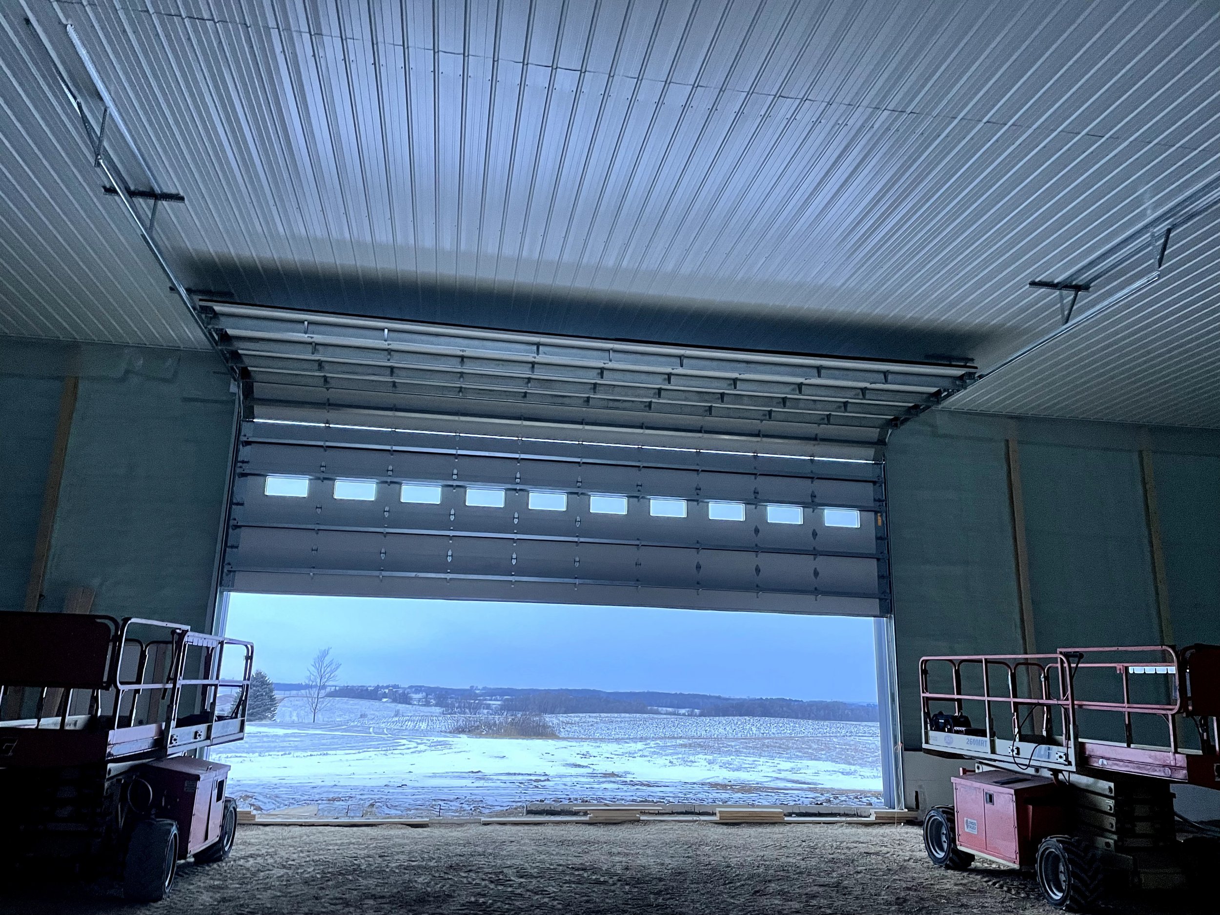 Open garage door revealing snow-covered field and trees outside, with farm equipment inside