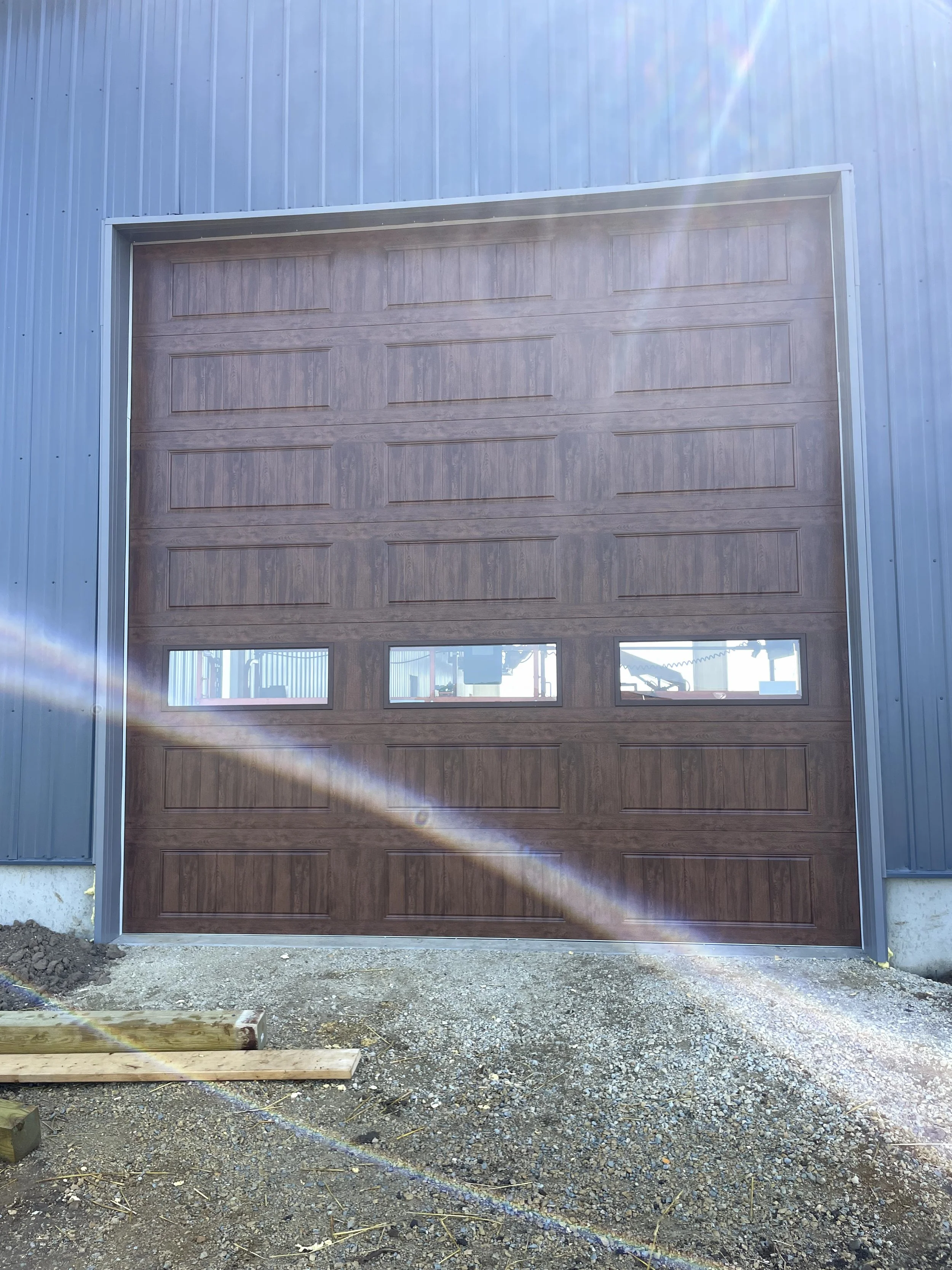 A closed brown garage door with four windows at the bottom, set in a metal building, with an unfinished gravel ground and wooden planks in front.