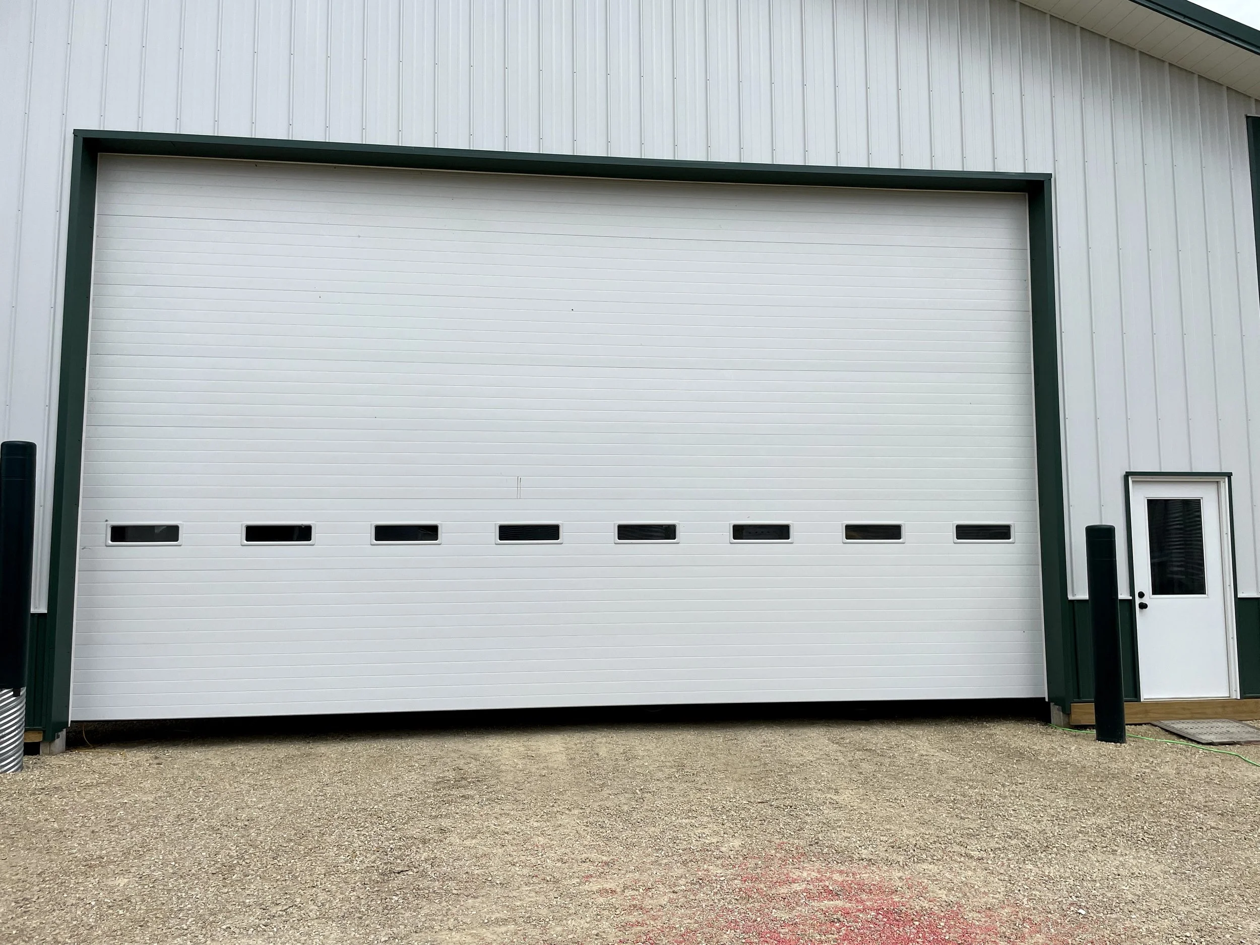 A large white industrial garage door on a metal building with small windows near the bottom, side steps, and black bollards in front on a gravel surface.