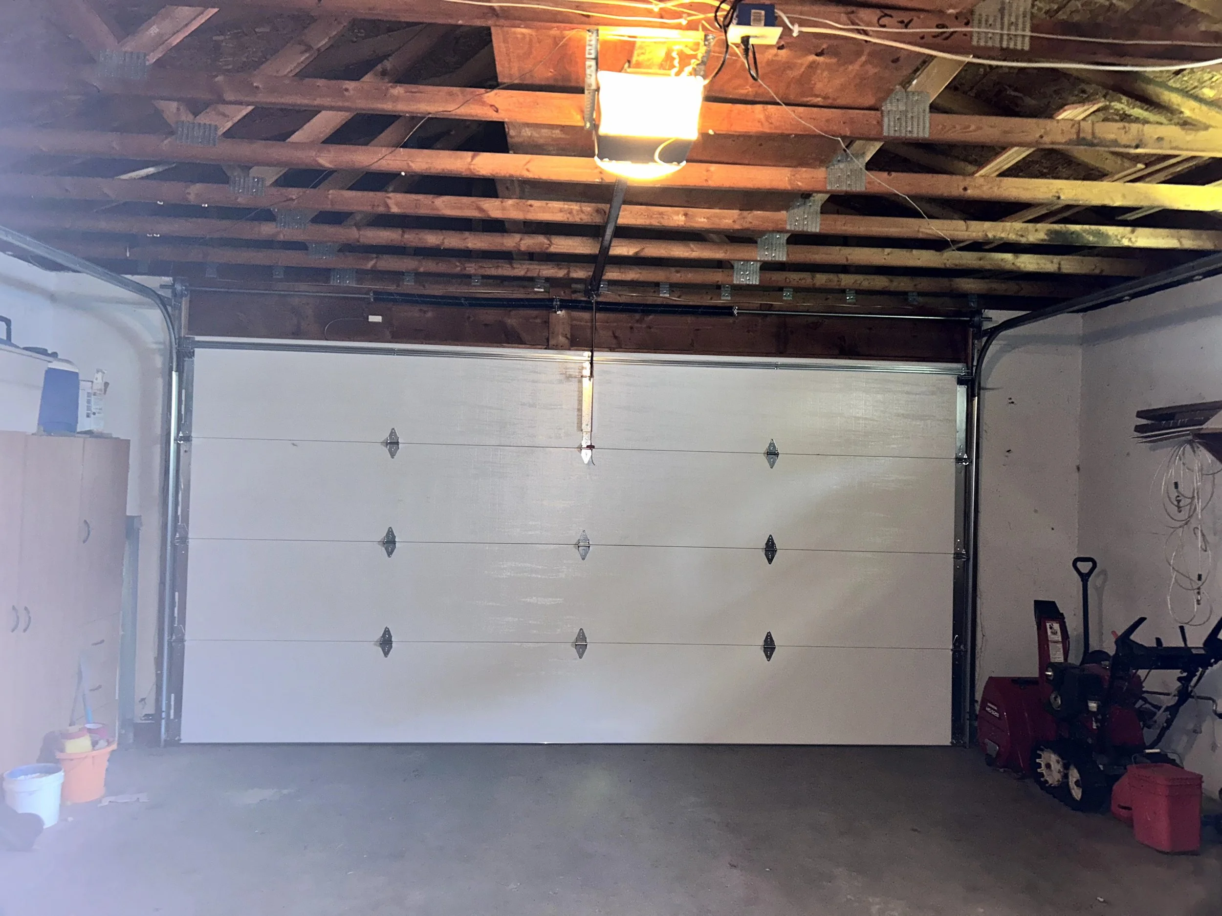 An unfinished garage interior with an open garage door, exposed wooden ceiling beams, and various tools and items on the concrete floor, including a red toolbox, a trash can, and a power drill.
