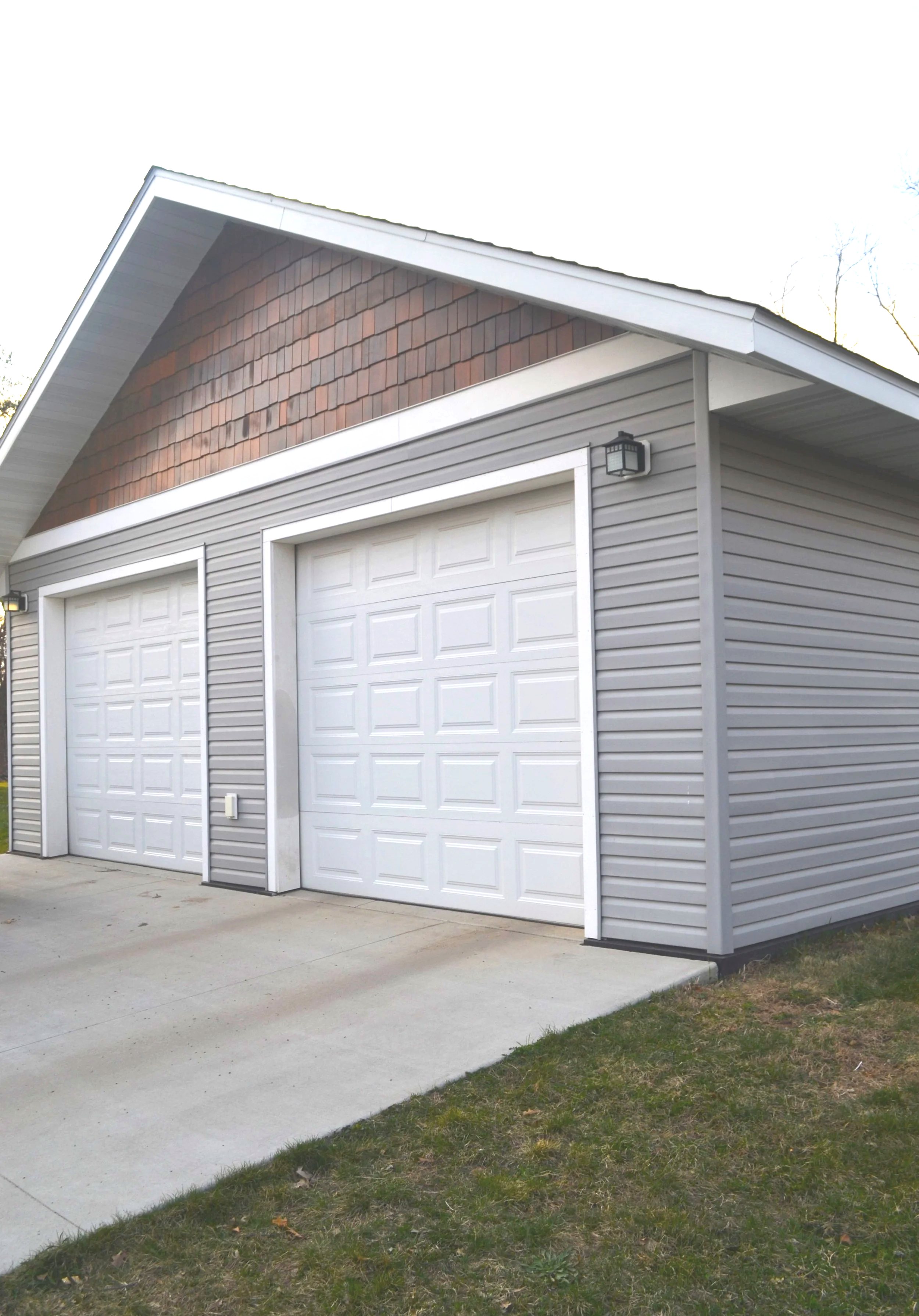A two-car garage with grey siding and white garage doors, a concrete driveway, and a grass lawn. The garage has a small outdoor light fixture on the right side.