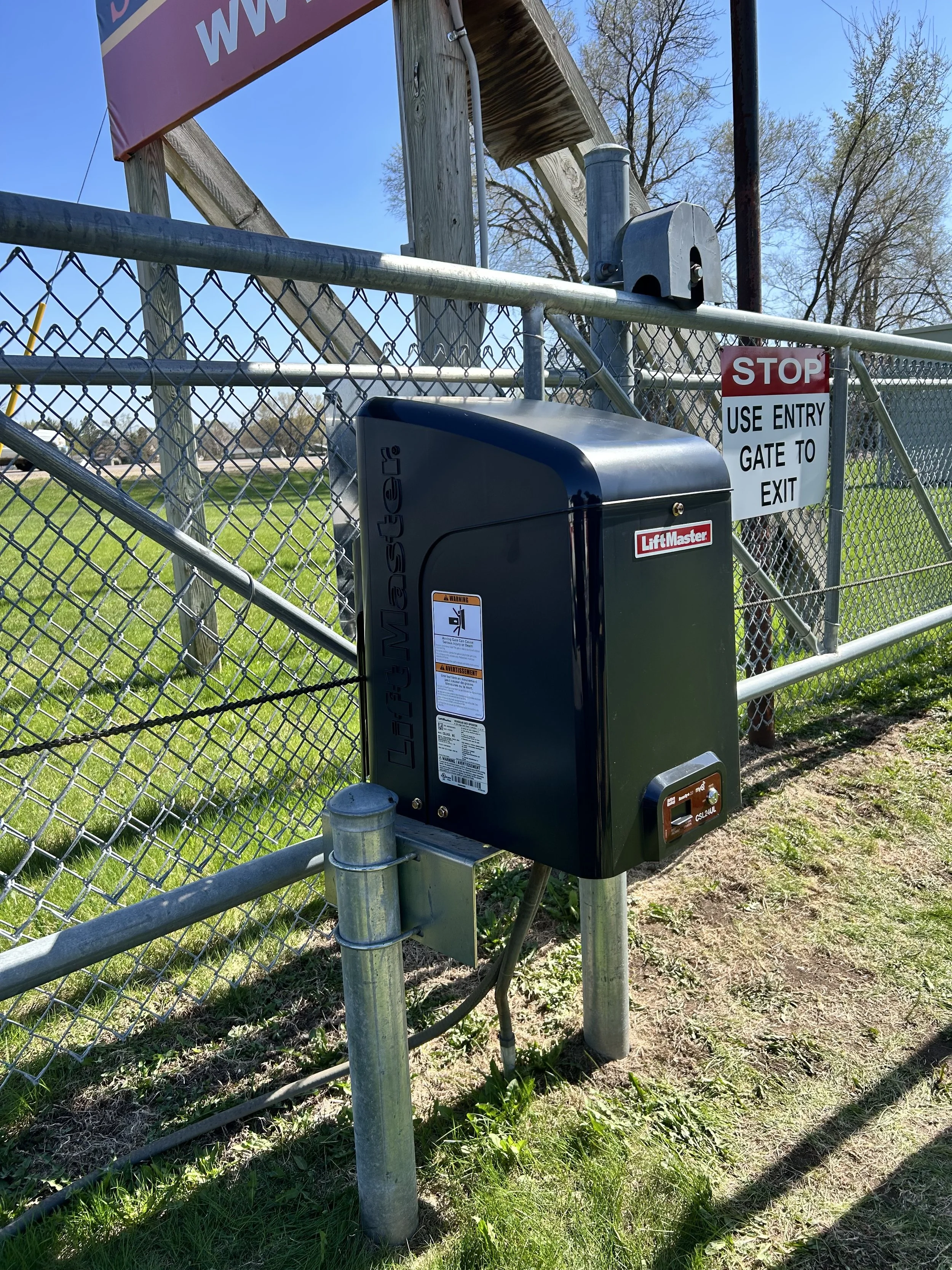 Close-up of a metallic gate control box with a warning label, attached to a chain-link fence, and a sign indicating entry and exit rules. The background shows trees and a clear blue sky.
