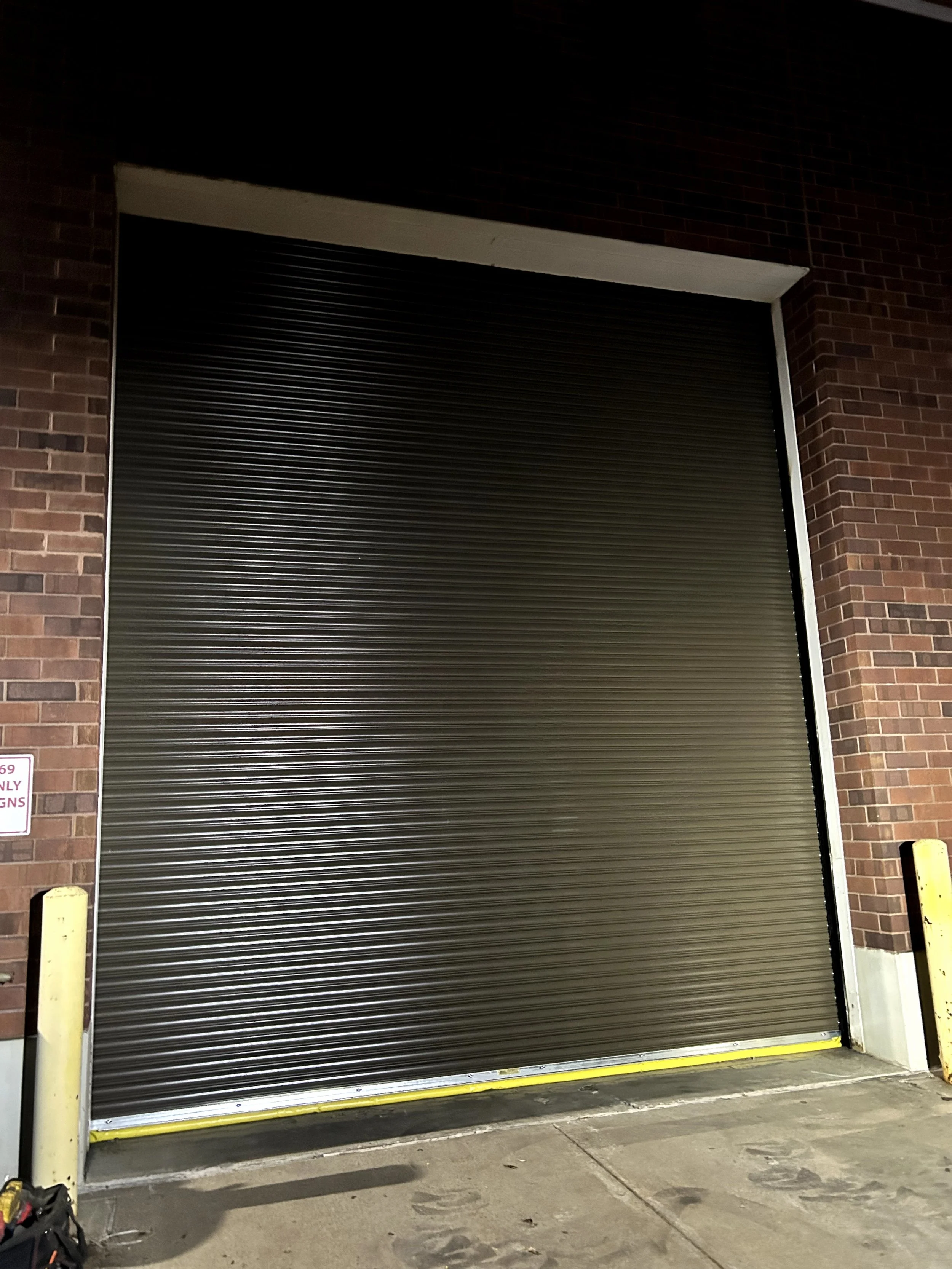 A closed, black garage door set in a brick wall at night, with yellow protective barriers on each side and a yellow strip at the bottom edge.