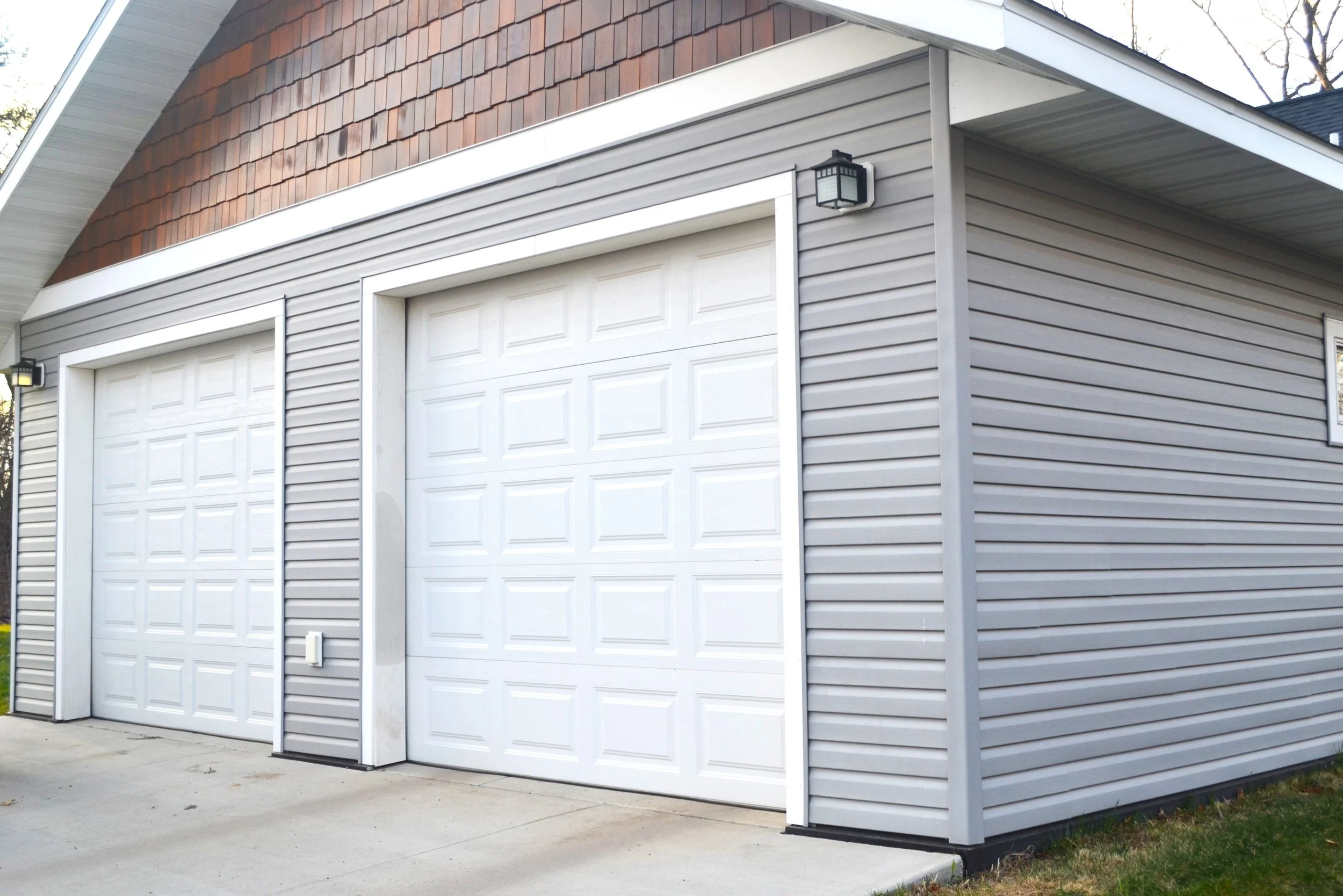 A residential garage with two white sectional doors, surrounded by grey vinyl siding, and a small outdoor light fixture mounted on the wall.