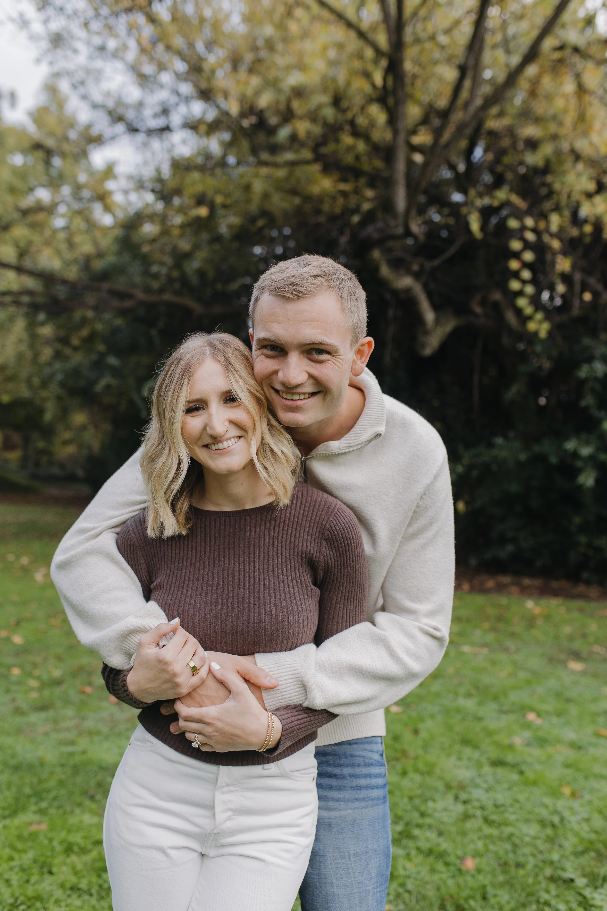 A smiling young woman with blonde hair, wearing a brown sweater and white pants, is being embraced from behind by a young man with short blonde hair, wearing a white sweater and blue jeans, in a park with green grass and autumn trees.