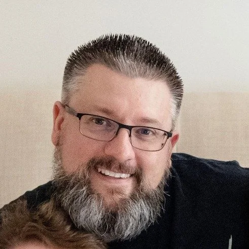 A smiling man with glasses and a beard, wearing a black shirt, in front of a beige background.