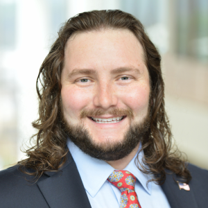 A man with long brown hair, a beard, wearing a dark suit, light blue shirt, and patterned red tie, smiling in an indoor setting