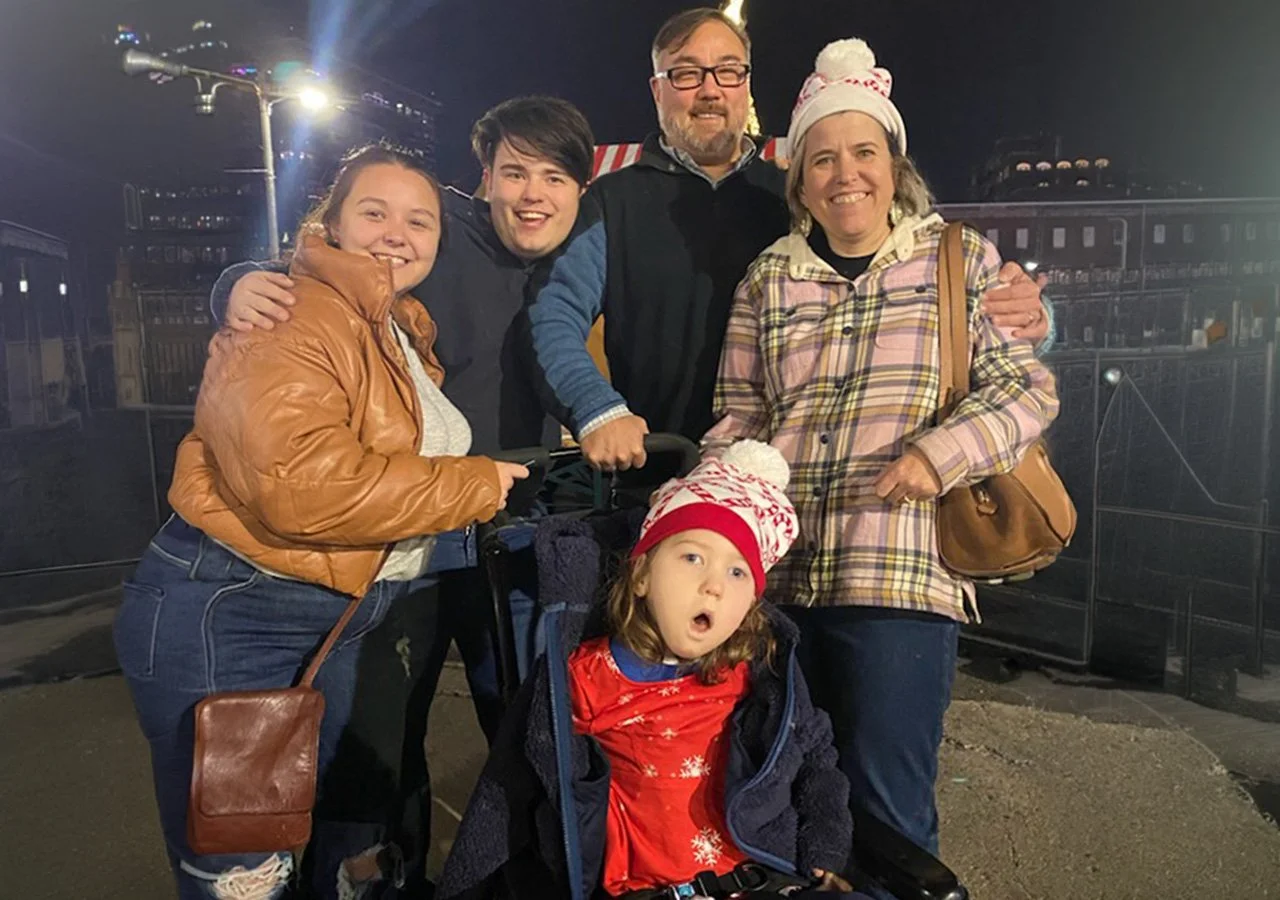 Group of six people, including a young girl in a stroller, smiling and posing together at night in an outdoor setting with festive lights and structures in the background, some wearing winter clothing and holiday accessories.