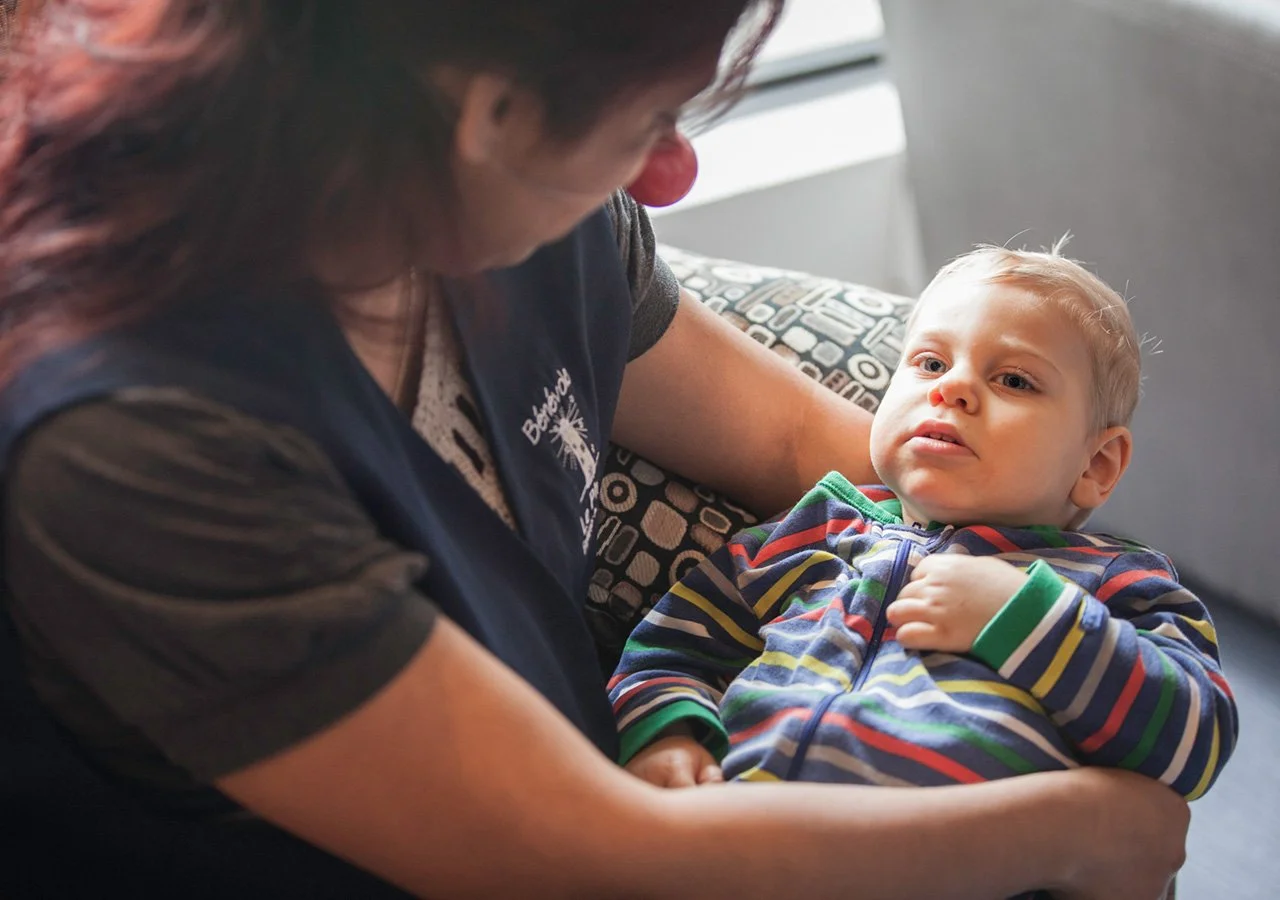 A woman with medium-length dark hair looking at a young boy with blonde hair, wearing a rainbow striped hoodie, as he sits in her lap on a patterned couch.