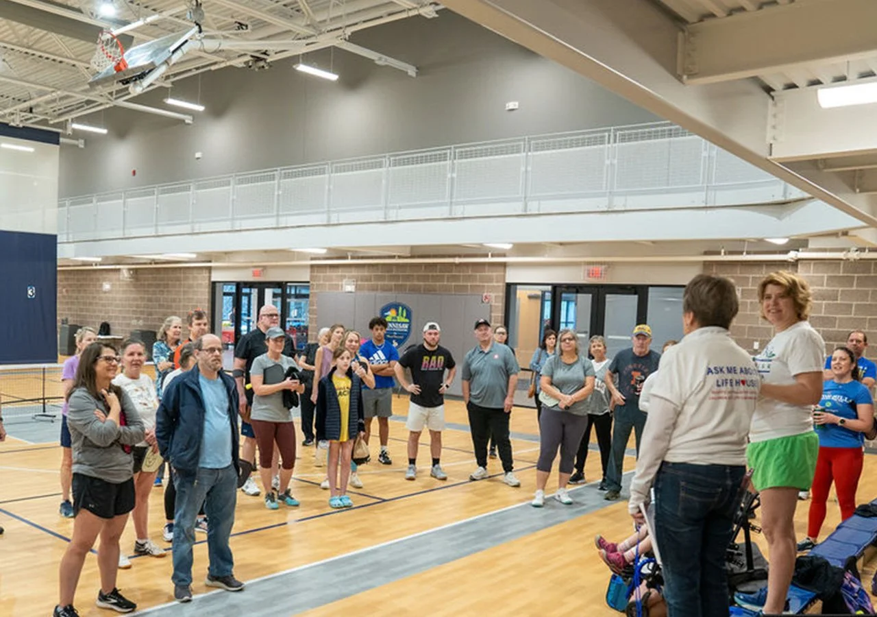 A group of people gathered in a gymnasium, listening to two women speaking at the front, with some holding clipboards, in front of a sign that says 'Lifeguard Pool'.