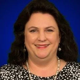 A portrait of a woman with dark hair, smiling, wearing a black and white patterned top and pearl necklace, against a blue background.