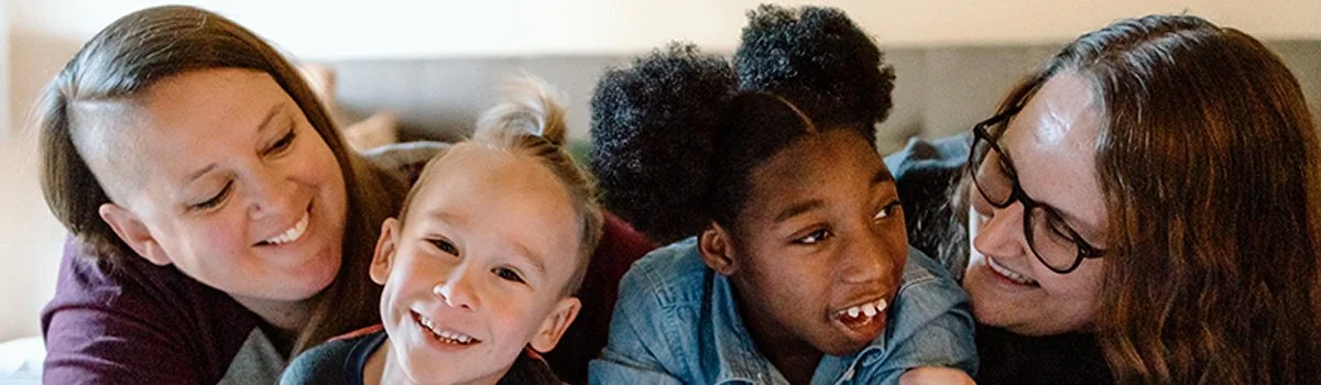 Two women and two kids smiling and lying close together indoors.