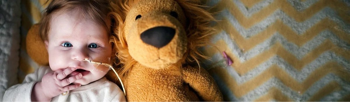 Close-up of a baby with blue eyes lying next to a stuffed lion toy, wearing a nasal cannula, on a zigzag patterned blanket.