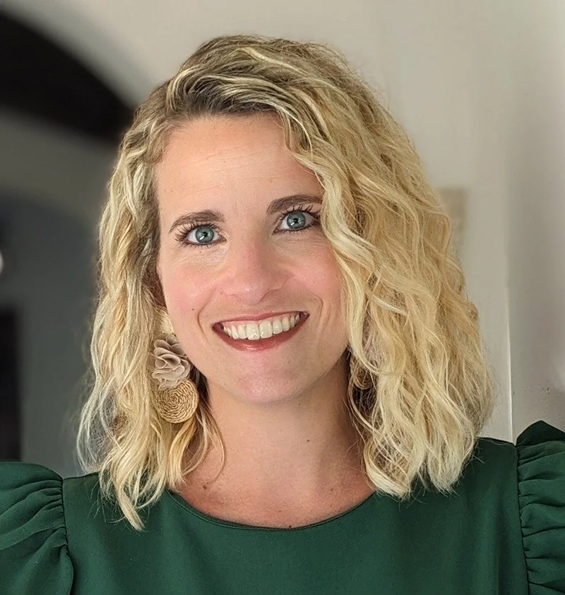 A woman with blonde, curly hair and blue eyes smiling, wearing a dark green top and floral earrings, standing indoors.