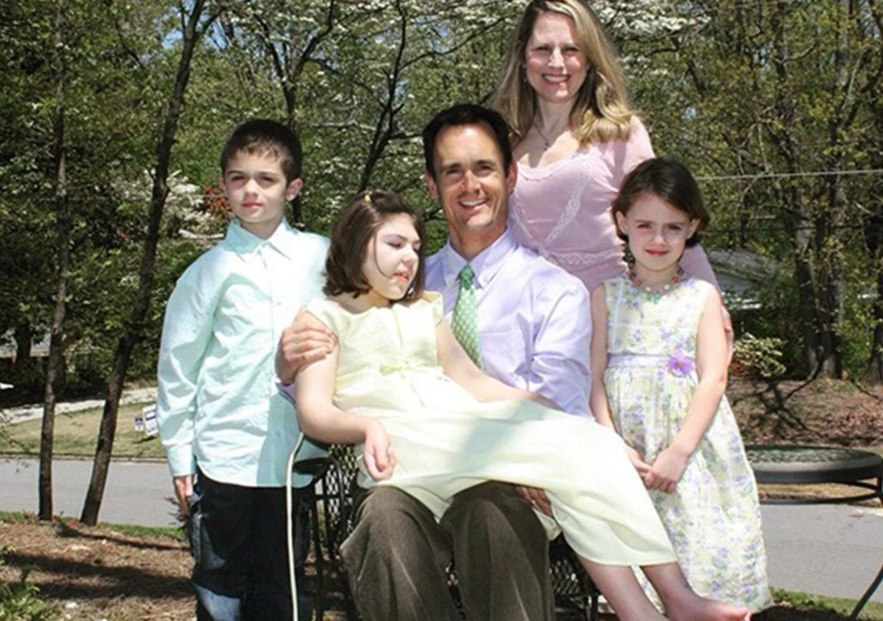 A family of six outdoors on a sunny day, sitting and standing around a wheelchair, with trees and a park in the background. The father is sitting in the wheelchair with a girl on his lap, surrounded by two boys and two women.