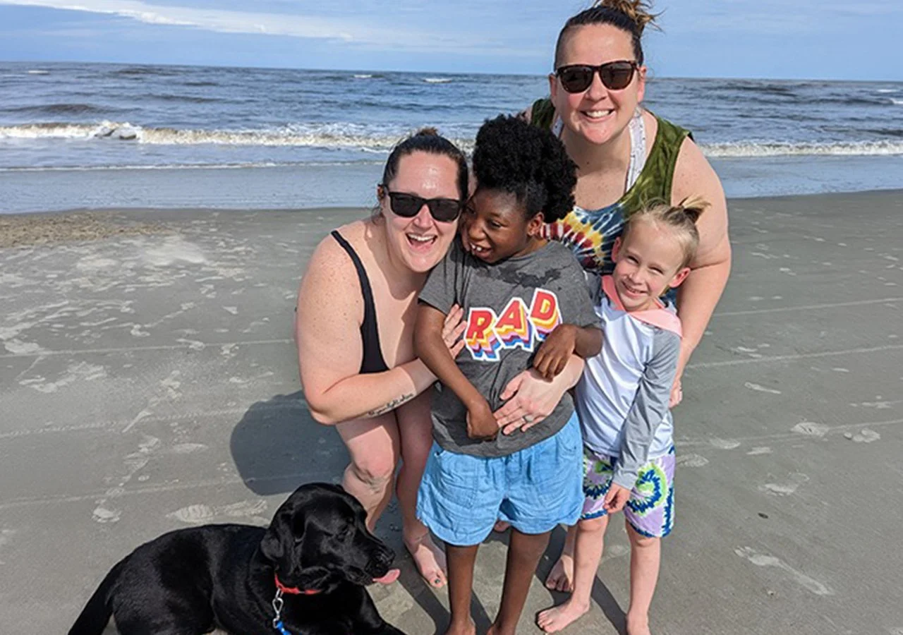 Four women and two children smiling on a beach, with a black dog sitting in front. The ocean and sky are in the background.