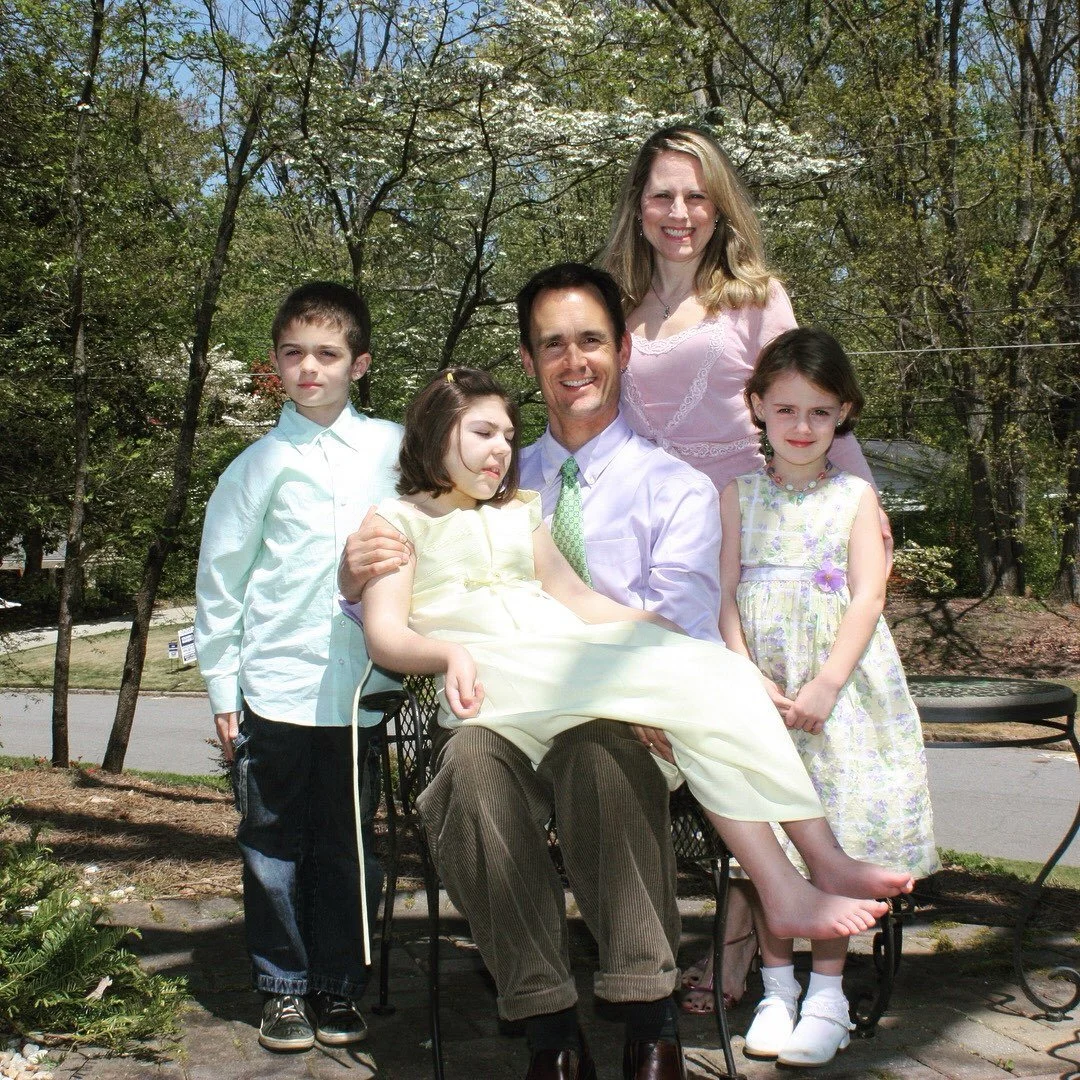 A family photo outdoors featuring a man, woman, and four children, with trees and blooming flowers in the background, enjoying a sunny day.