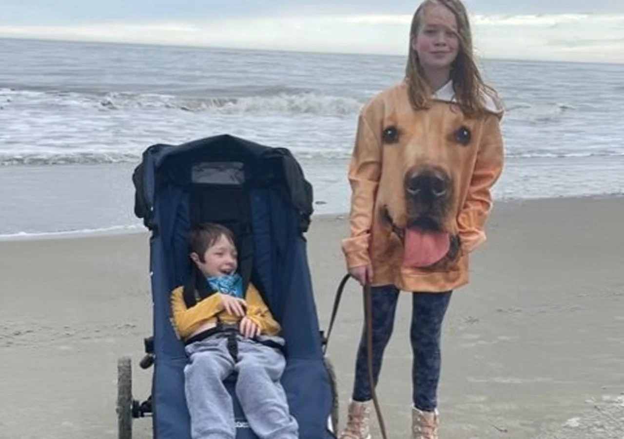 A girl and a boy on a beach; the girl wears a jacket with a large dog face print, and the boy sits in a stroller, smiling.