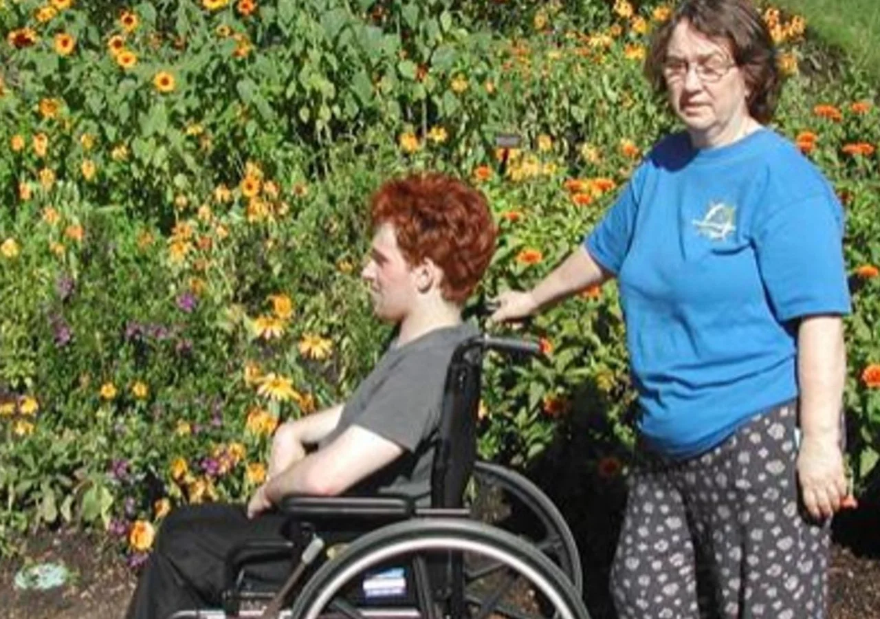A woman with red hair sitting in a wheelchair next to a woman with glasses and brown hair, wearing a blue t-shirt and patterned pajama pants, standing outdoors in front of a garden with yellow and orange flowers.