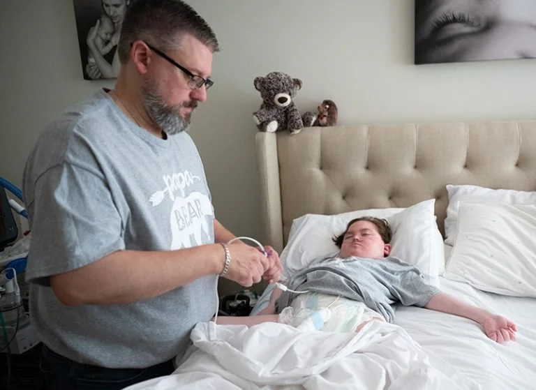A man in a gray t-shirt standing beside a hospital bed, listening to a young girl's heartbeat with a stethoscope. The girl is lying in bed, with her eyes closed, covered with a blanket. The room has a neutral wall with framed black and white photos and stuffed animals on the headboard.