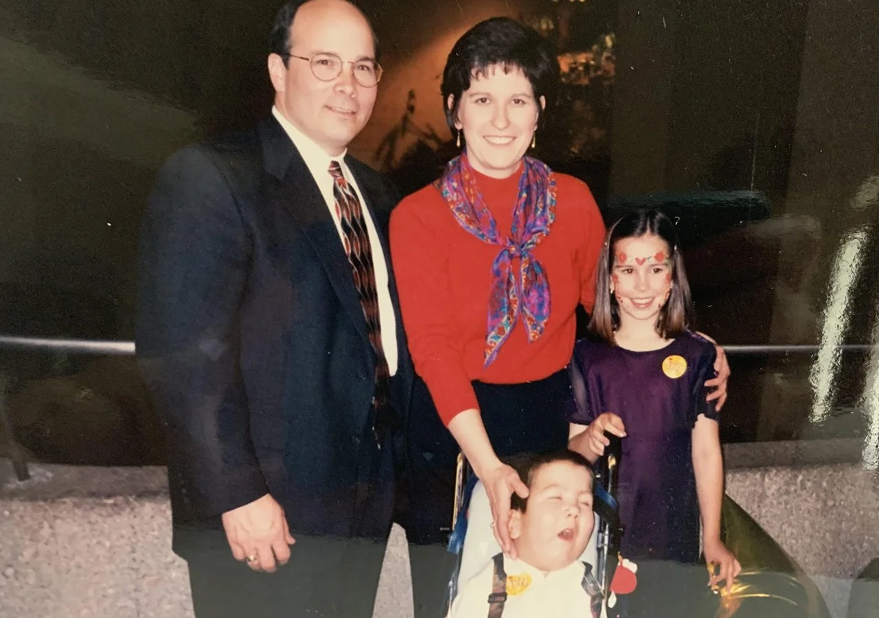 A family of four posing together indoors, including two adults and two children, one in a wheelchair.