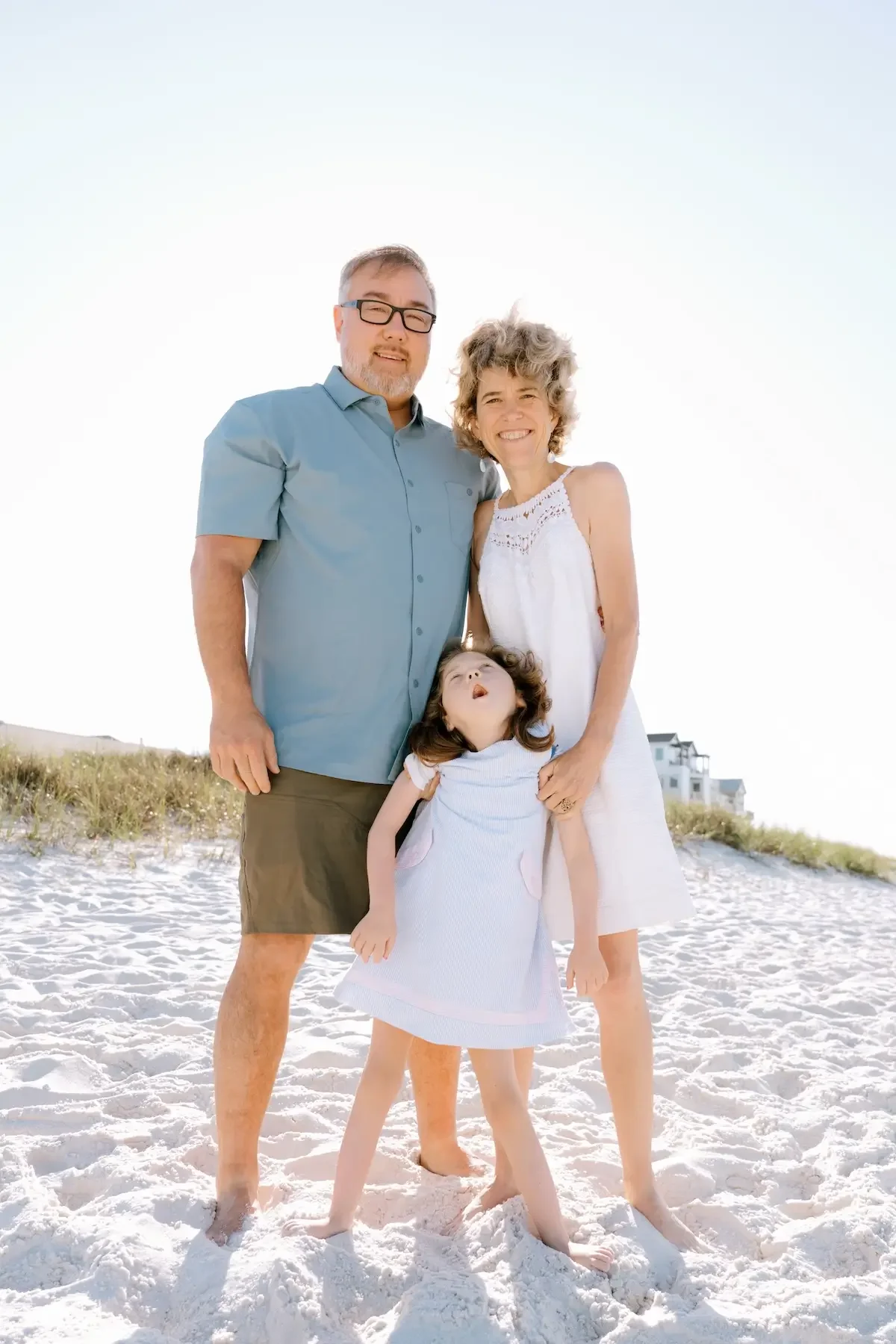 A family of four standing on a sandy beach near dunes, smiling, with clear sky and a house in the background.