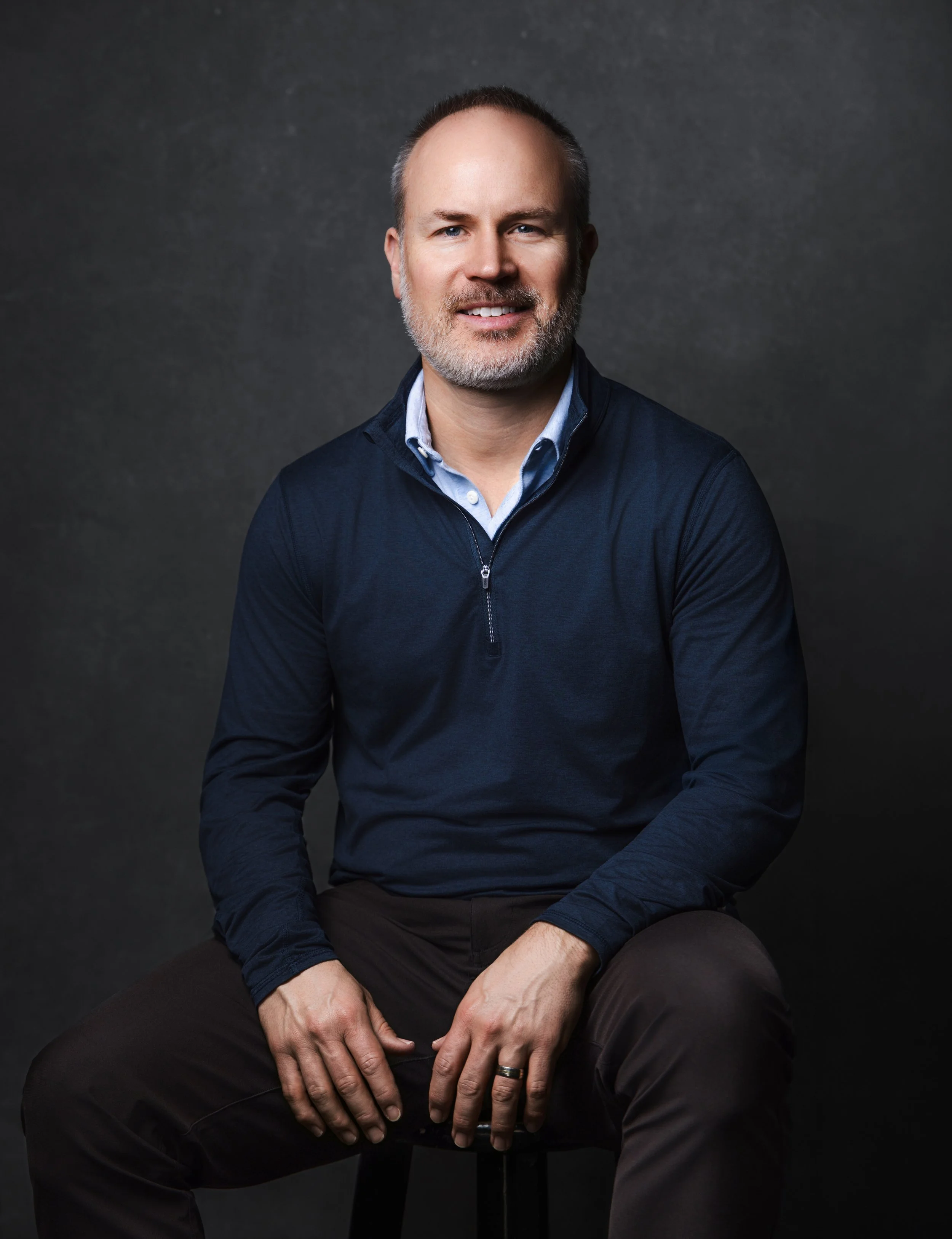 Campbell Gerrish sitting against a dark grey background, smiling slightly, wearing a blue zip-up sweater over a light blue collared shirt, with his hands resting on his lap.