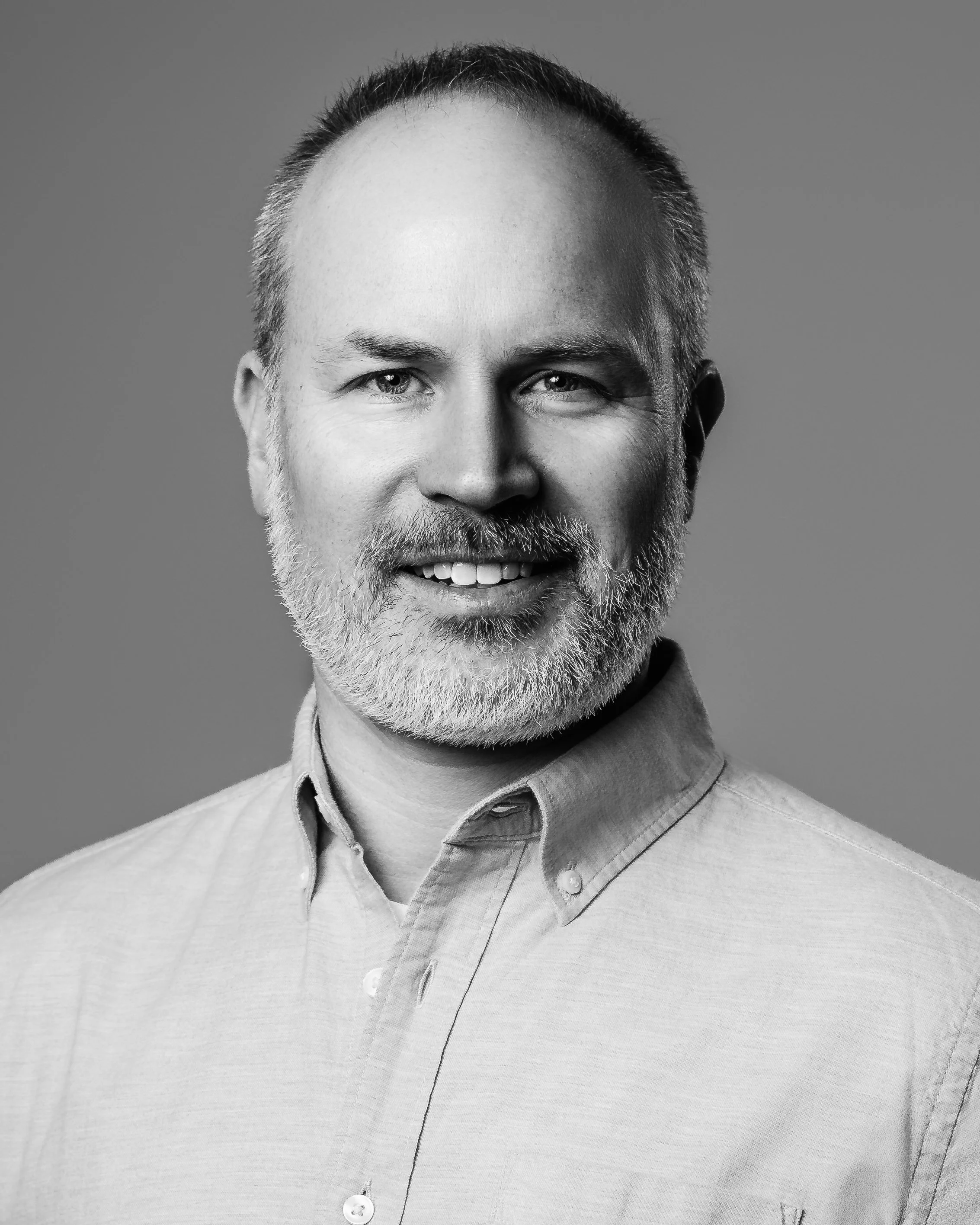 Black and white photo of a smiling Caucasian man with short hair and a beard, wearing a button-up shirt.