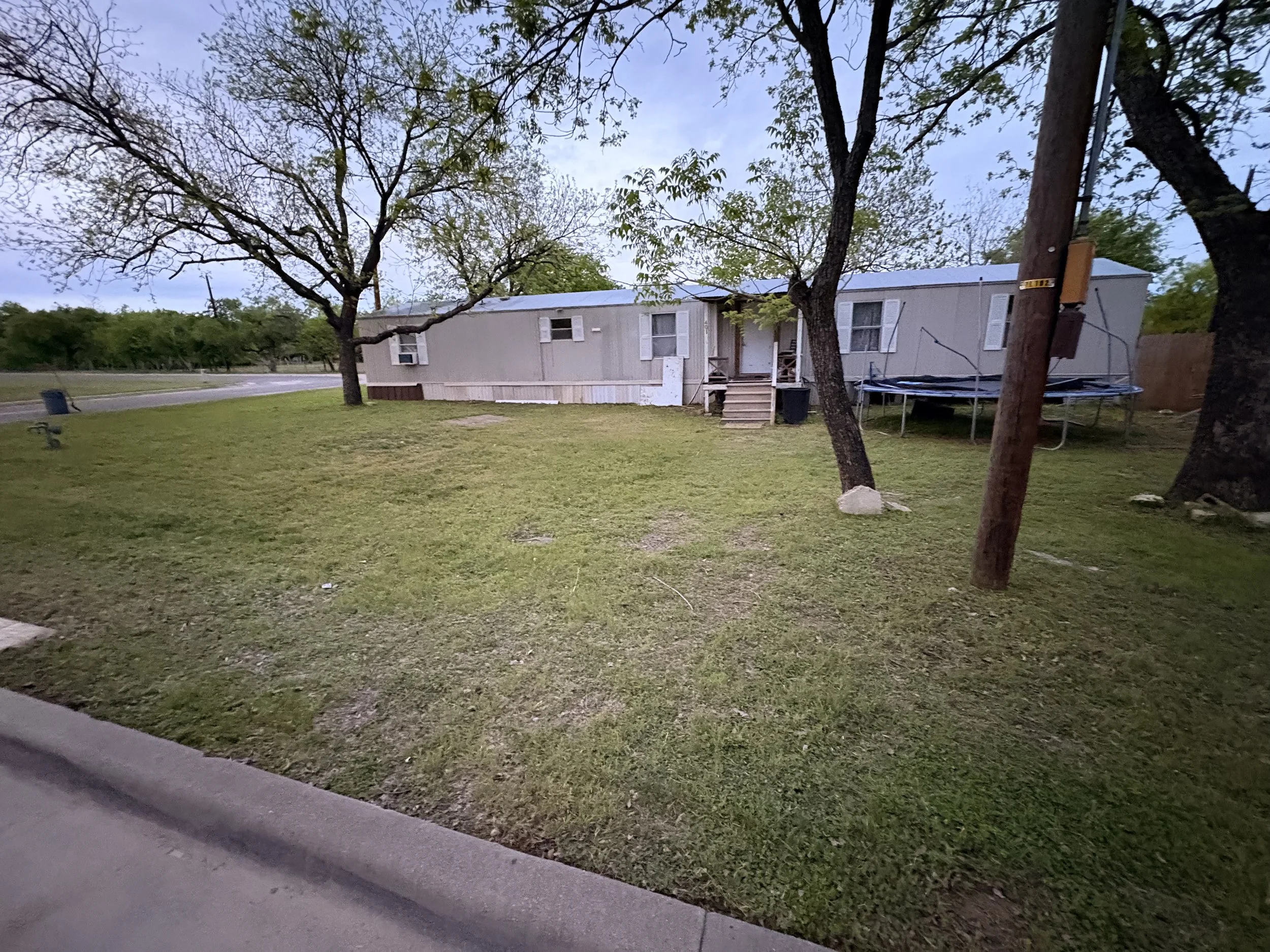 A mobile home situated in a grassy area with trees, a trampoline nearby, and an electric pole on the right side. The sky is partly cloudy.