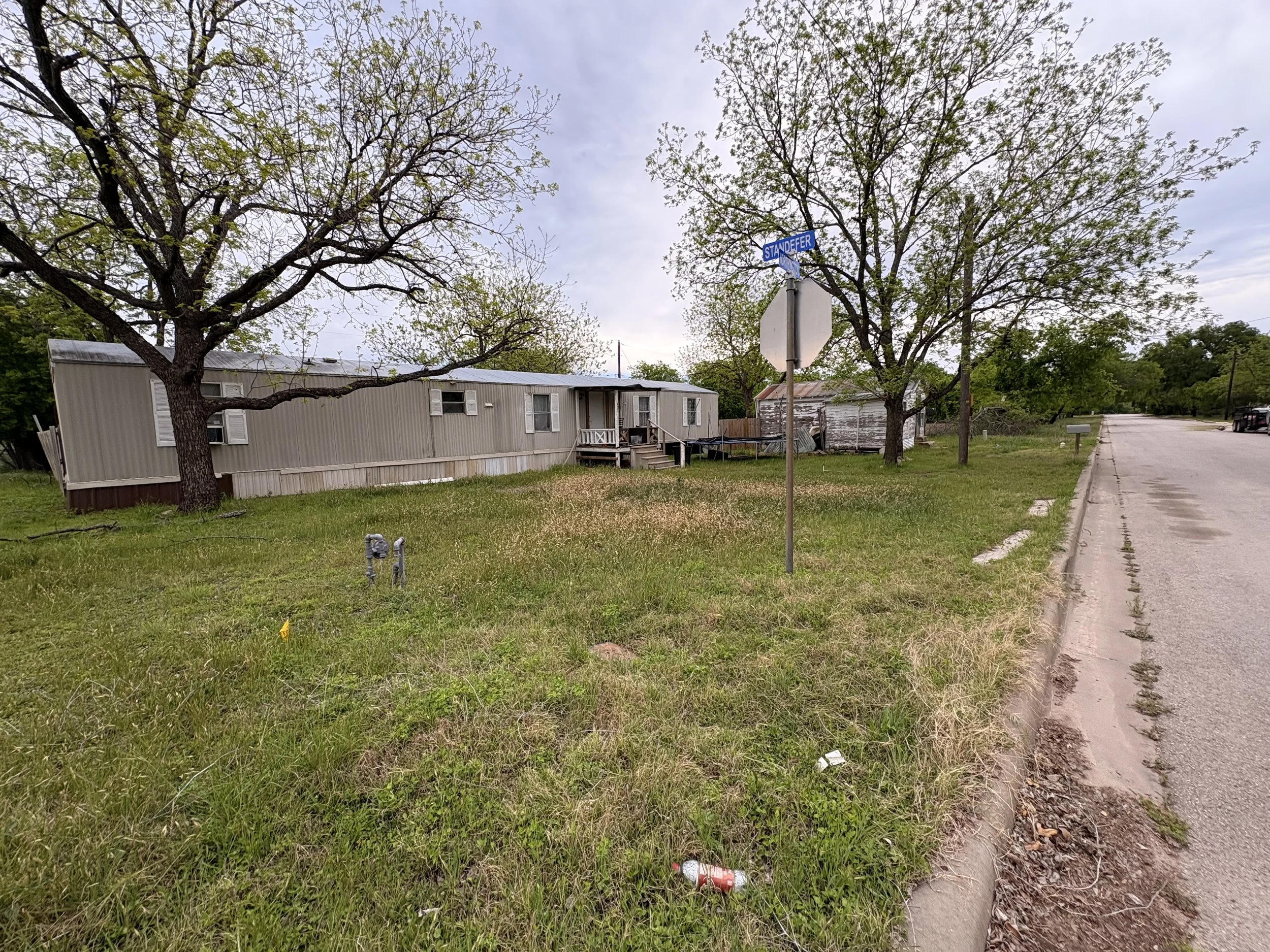 A mobile home on a grassy lot with two trees, a sidewalk, a street sign that reads 'Standefer,' and a small white building in the background. The sky is partly cloudy.