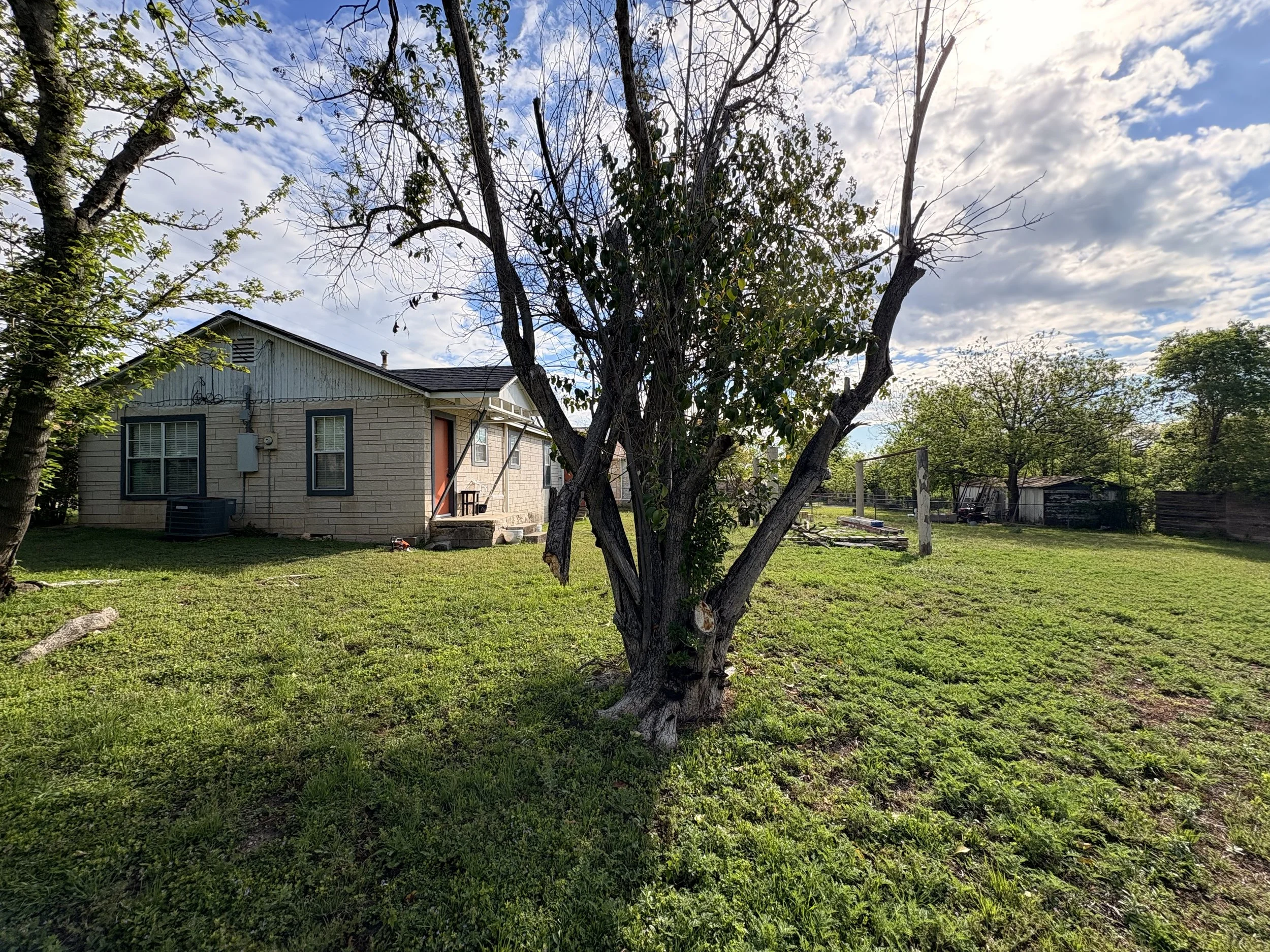 A backyard with a tree in the center, a small house with a brick exterior, a small porch, and a green lawn. There is a clothesline and several trees in the background, with a partly cloudy sky overhead.