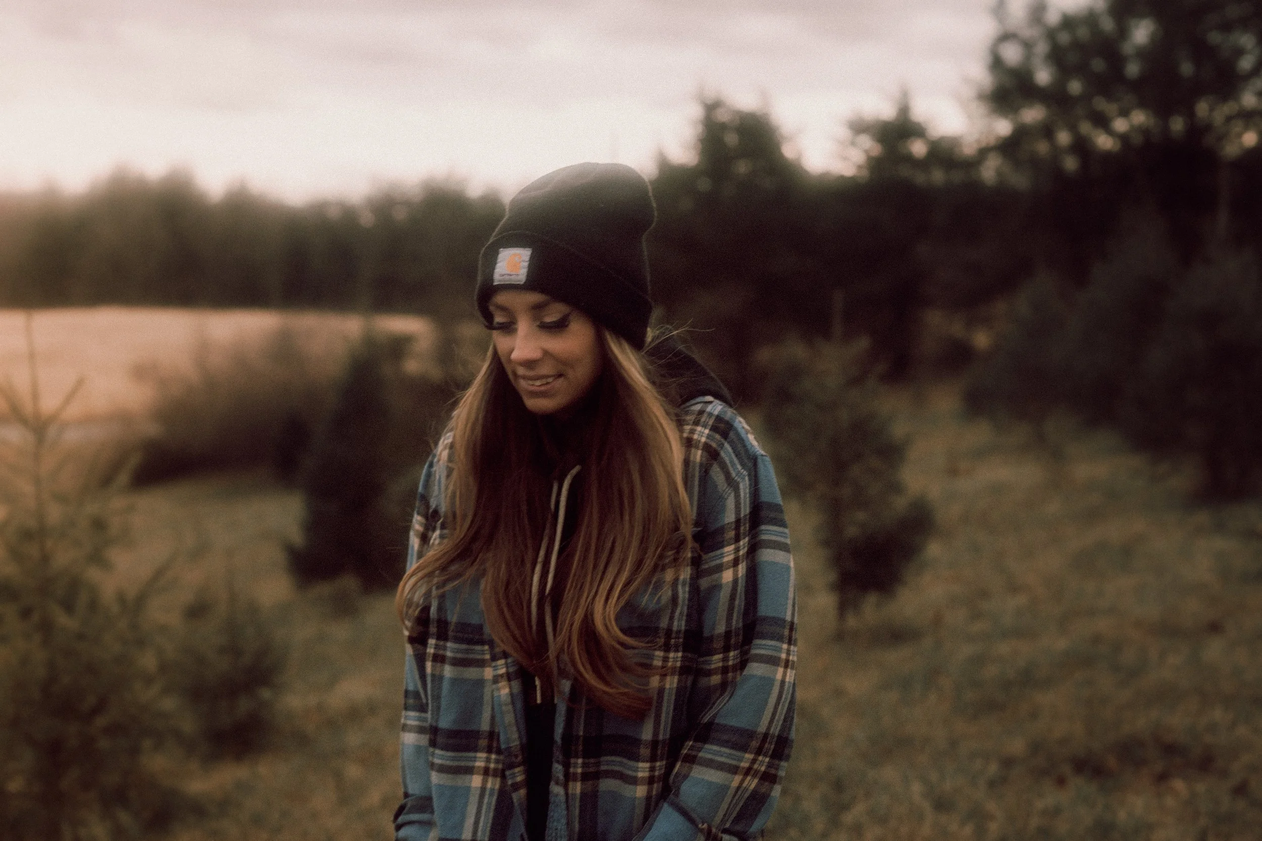 A young woman with long hair wearing a black beanie and a plaid shirt stands outdoors in a rural area during sunset with trees and fields in the background.