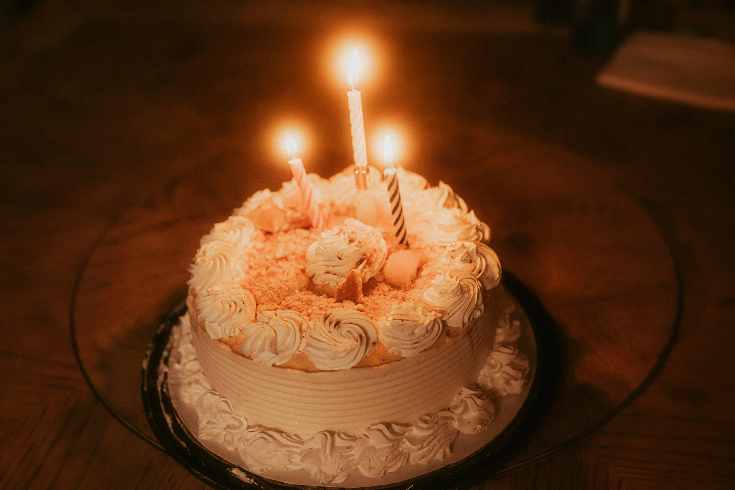 A birthday cake with three lit candles, decorated with cream swirls and a crumb topping, on a wooden table.