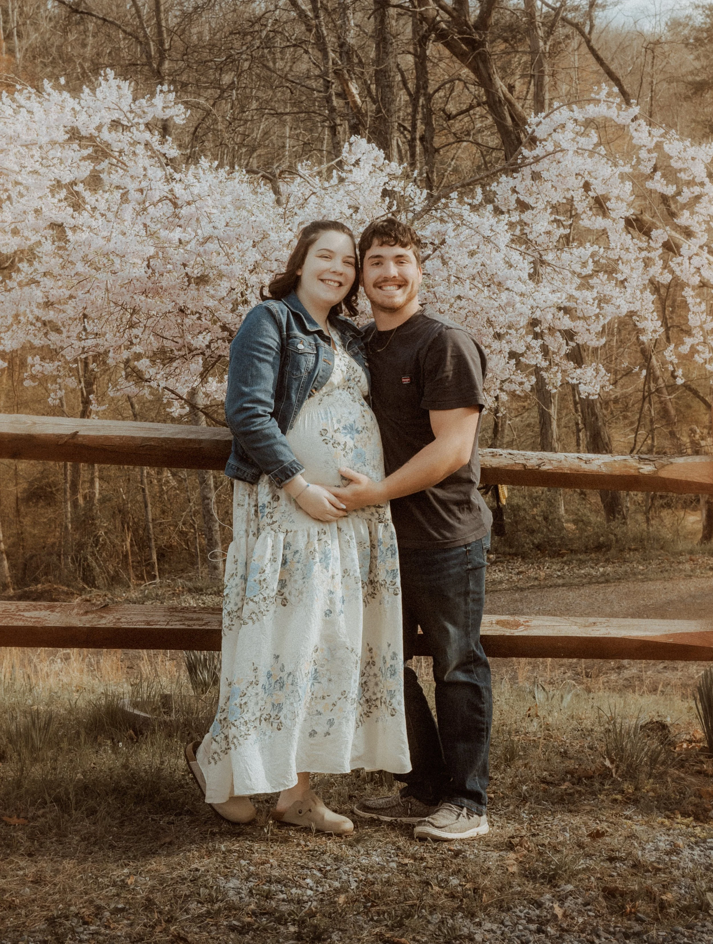 A pregnant woman and a man stand together outdoors in front of blooming trees, smiling and holding her belly.
