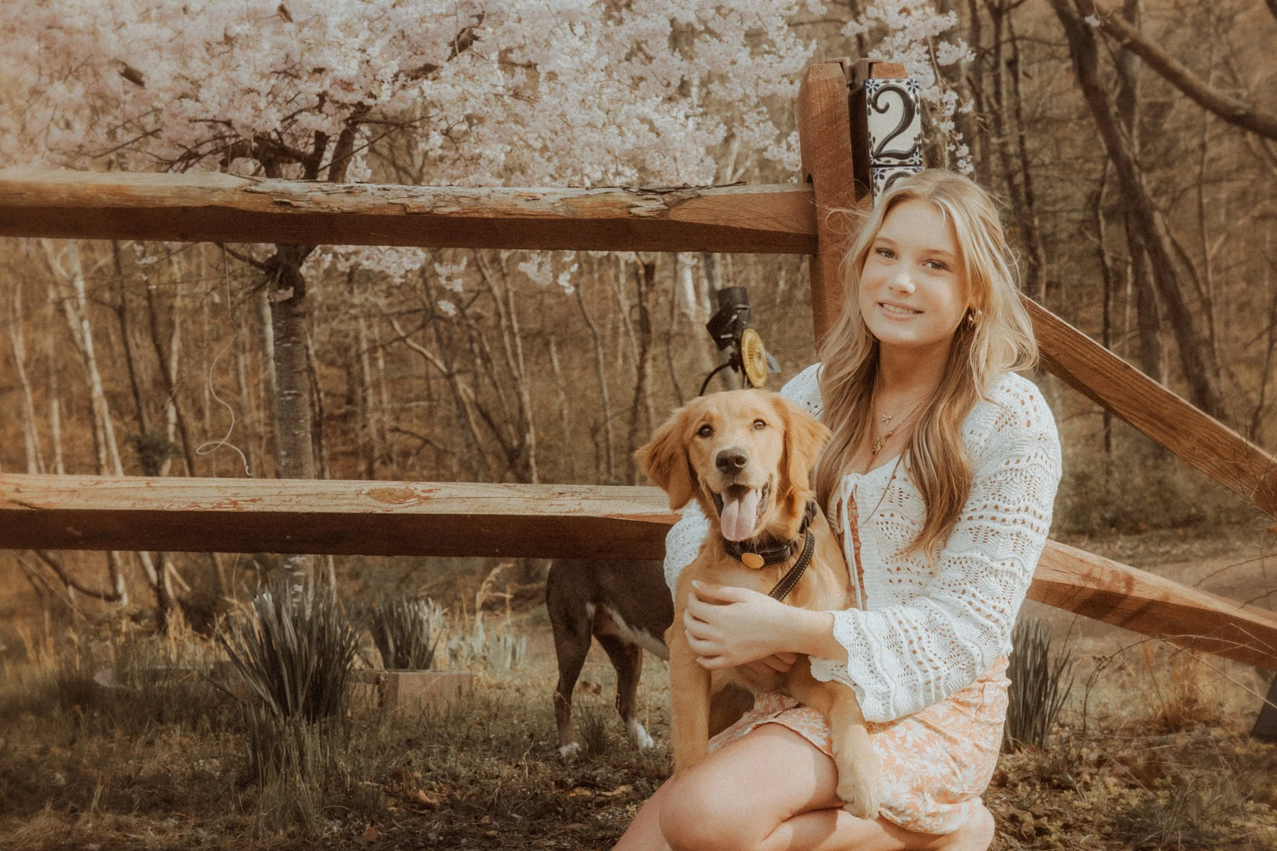 A young woman kneeling outdoors holding a golden retriever puppy, with wooded area and a wooden staircase in the background, during fall.