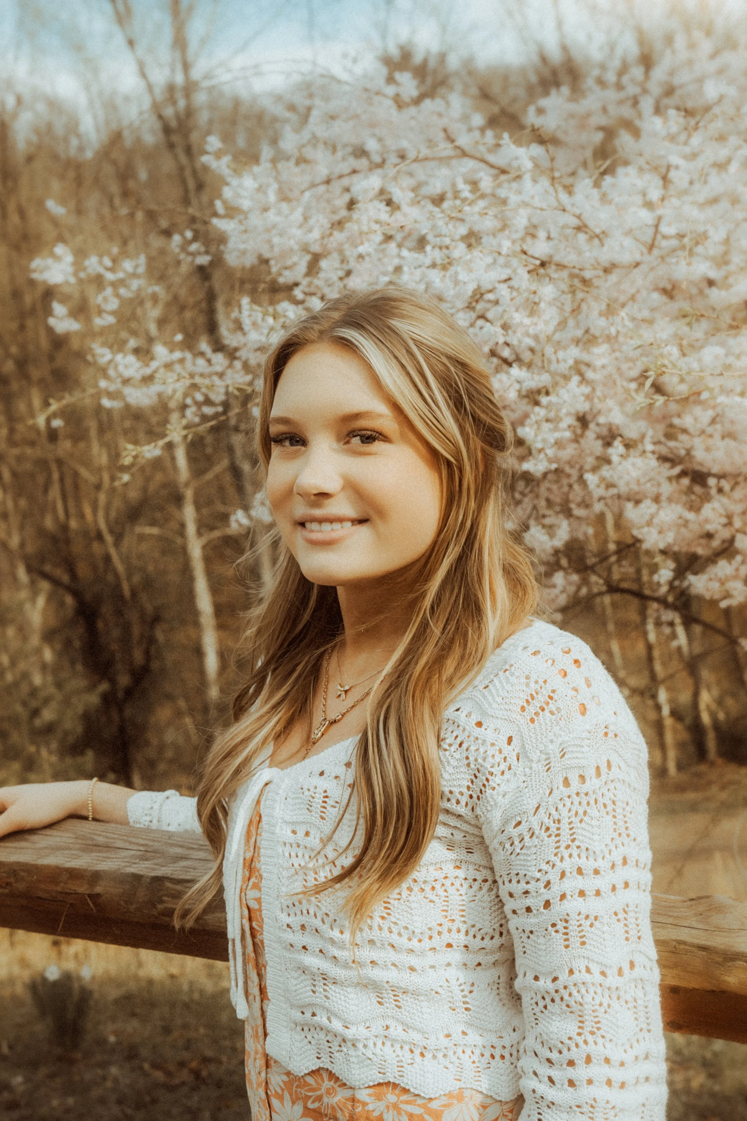 A young woman with long wavy hair, smiling, stands outdoors next to a wooden railing with blooming cherry blossoms in the background.