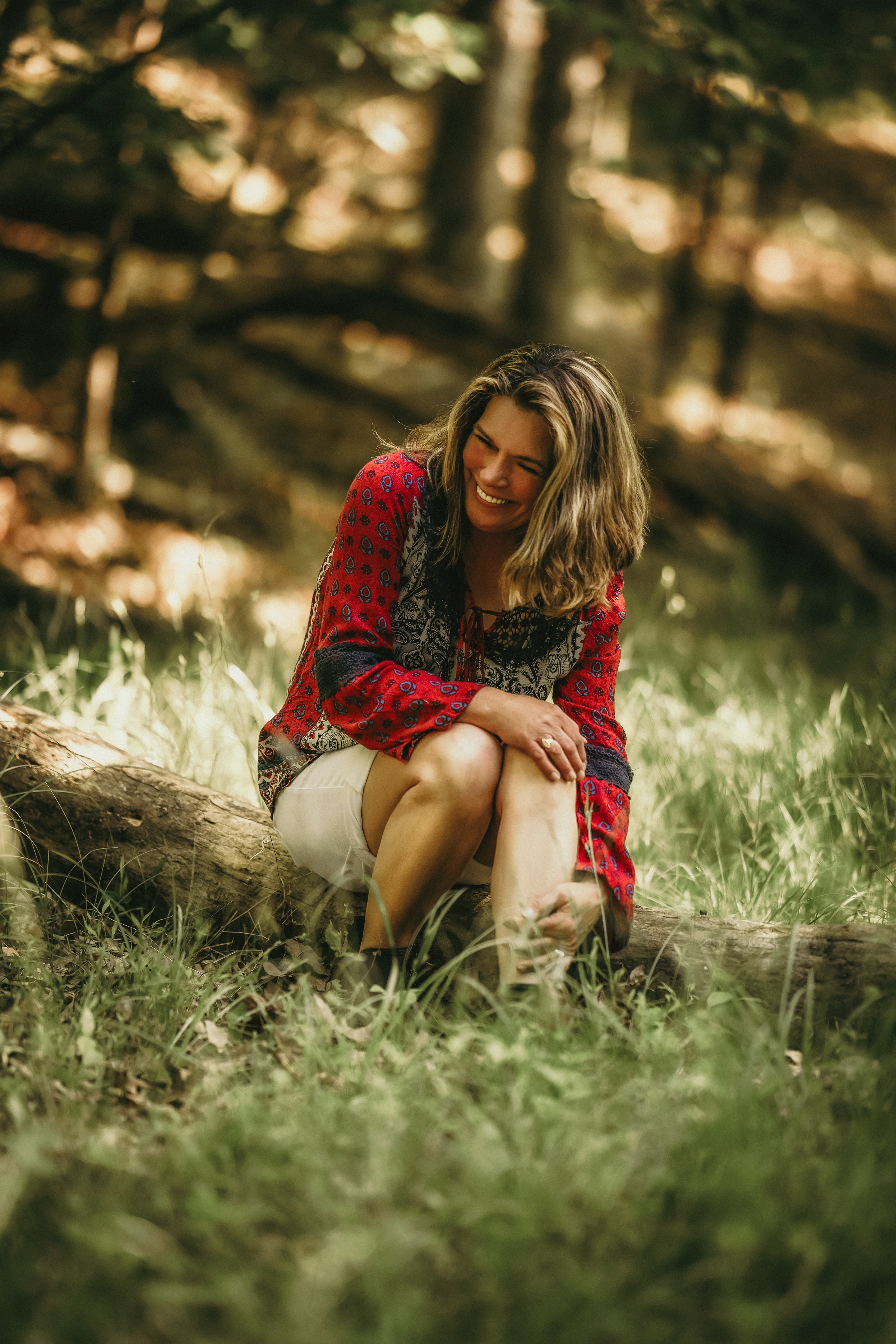 A woman sitting on a log in a forest, smiling and laughing.
